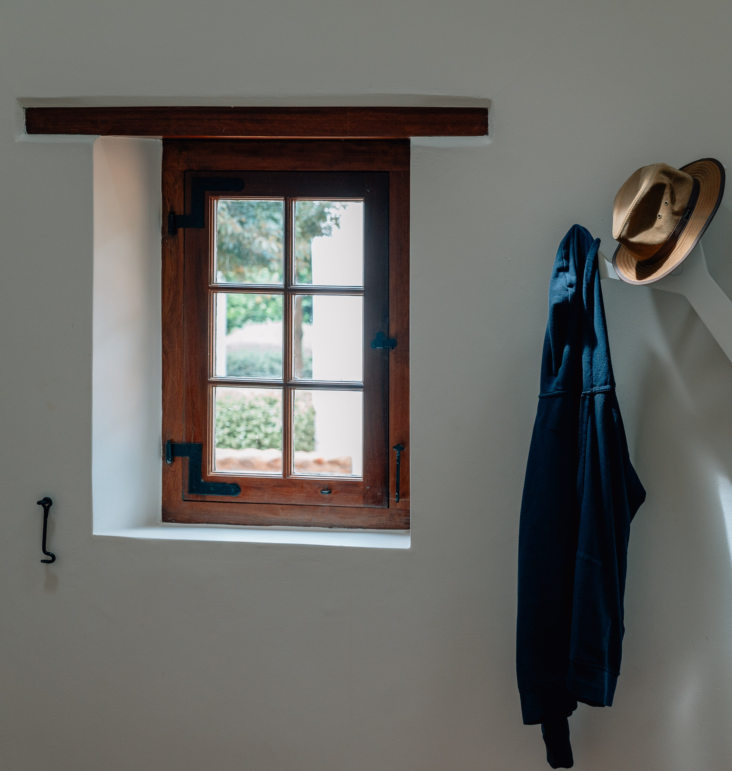 A rustic wooden-framed window with six panes overlooks a garden, while a tan cowboy hat and dark blue jacket hang on the pale gray wall beside it