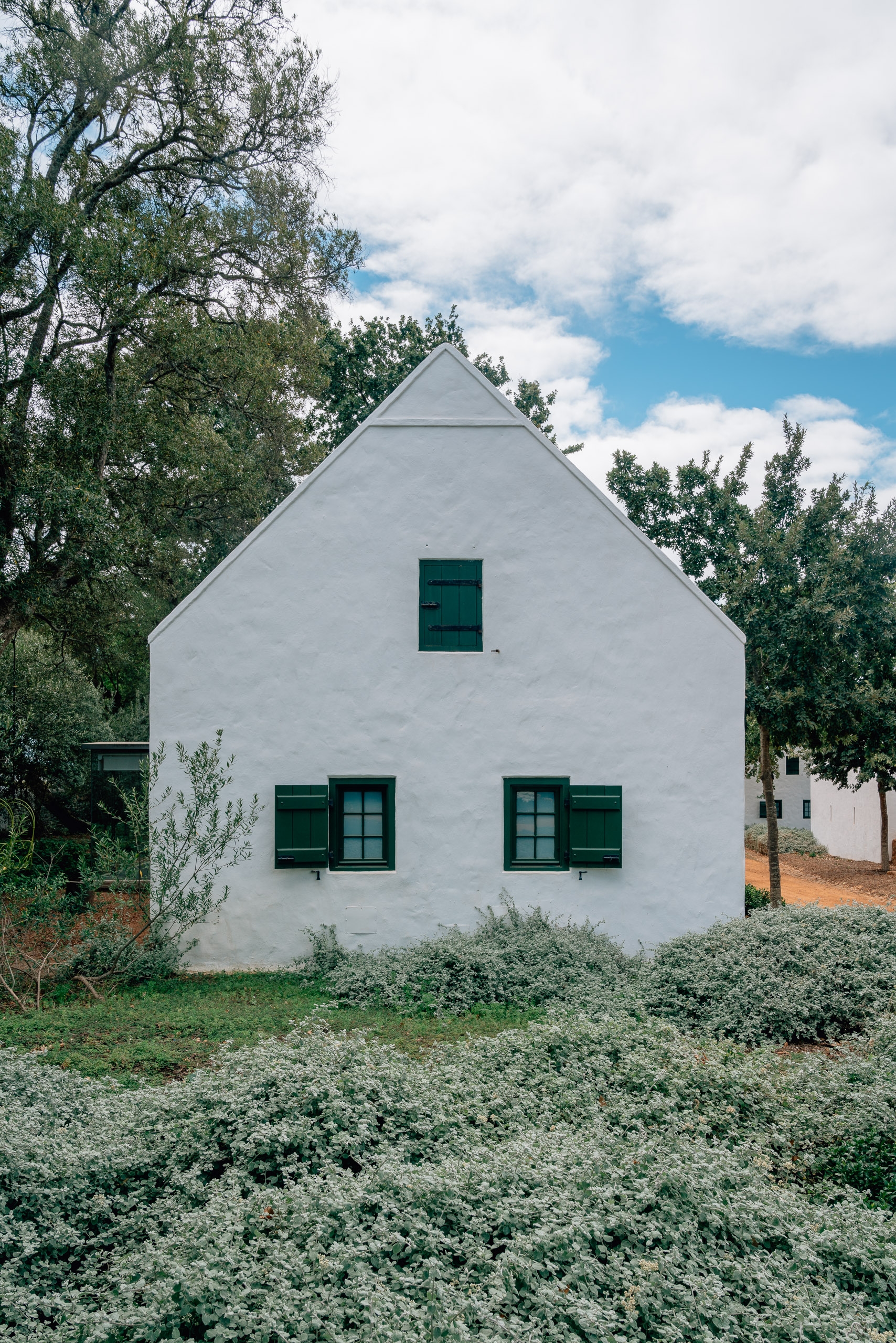 A whitewashed cottage with a peaked gable roof and dark green shuttered windows sits surrounded by silvery-green foliage and tall trees under a partly cloudy sky