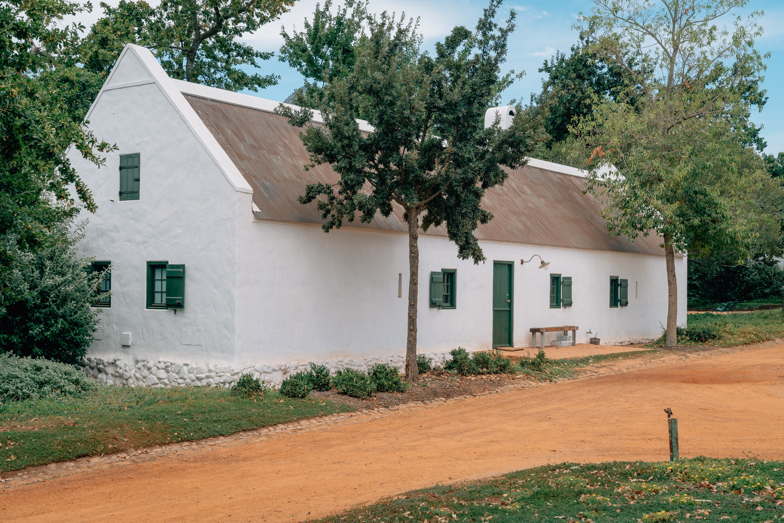 A whitewashed farmhouse with thatched roof and green shutters sits beside a red dirt road, surrounded by mature trees under clear blue sky.