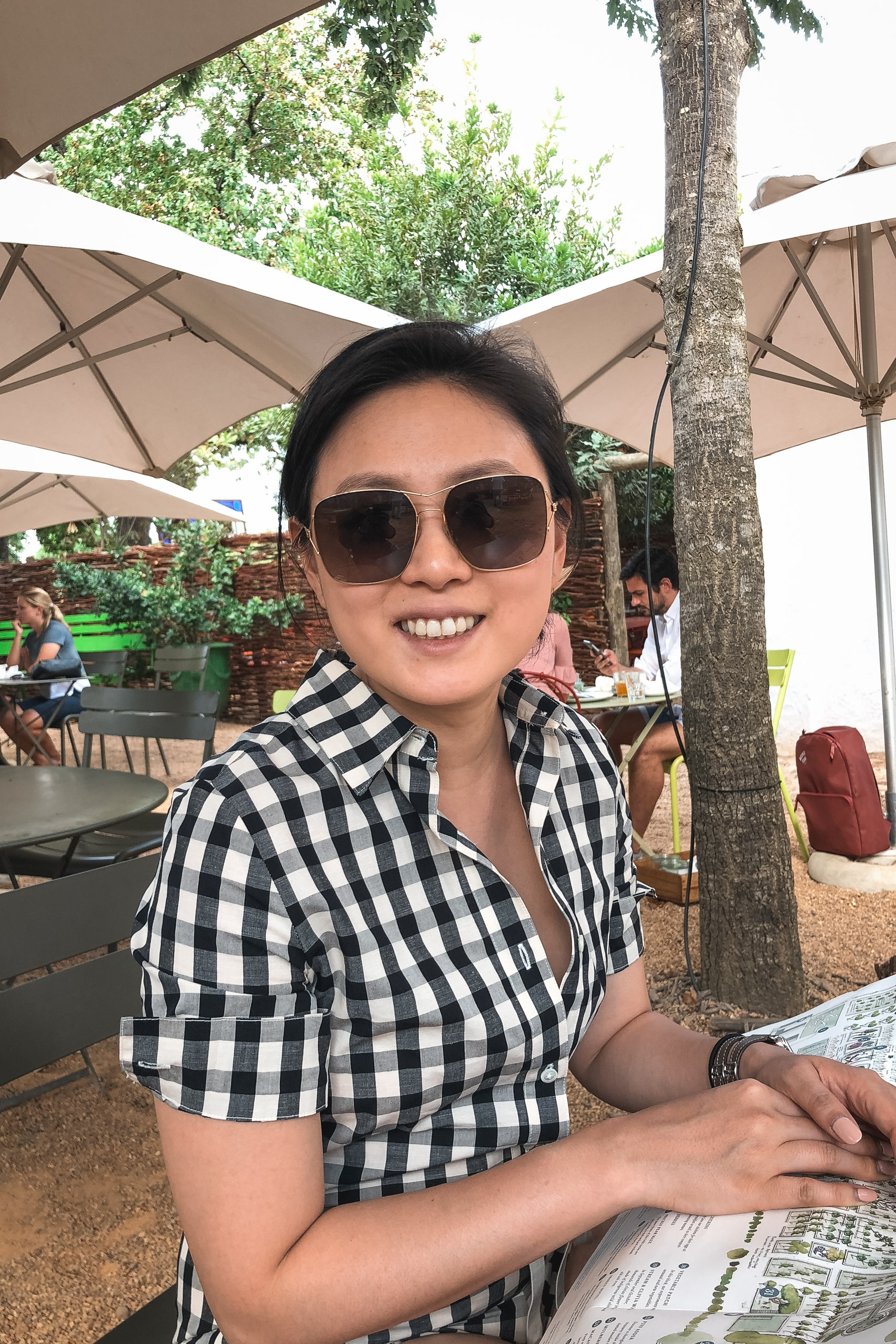 Woman in black and white checkered shirt and sunglasses smiles at outdoor patio shaded by umbrellas and palm trees, with other diners visible in background