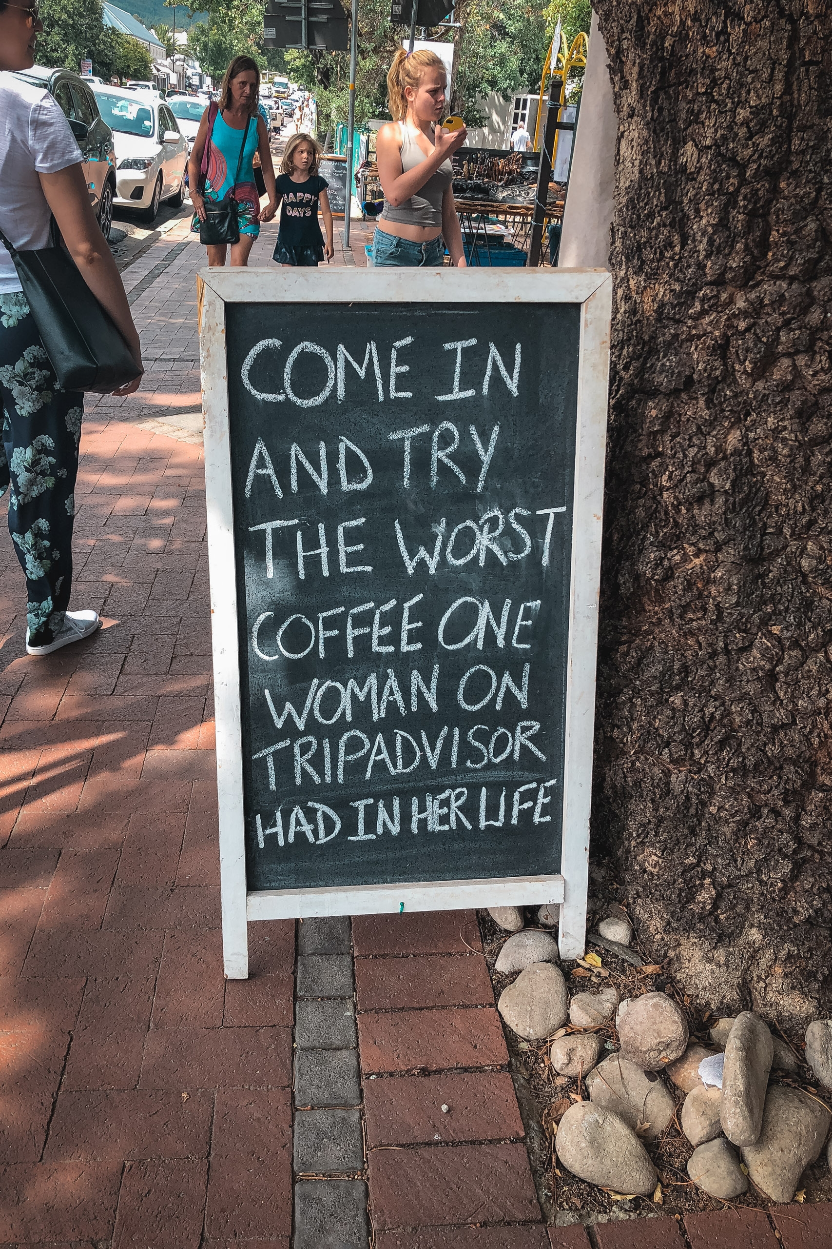 A chalkboard sandwich sign on a brick sidewalk displays humorous text inviting customers inside a coffee shop, with pedestrians and parked cars visible in the sunny street behind it.