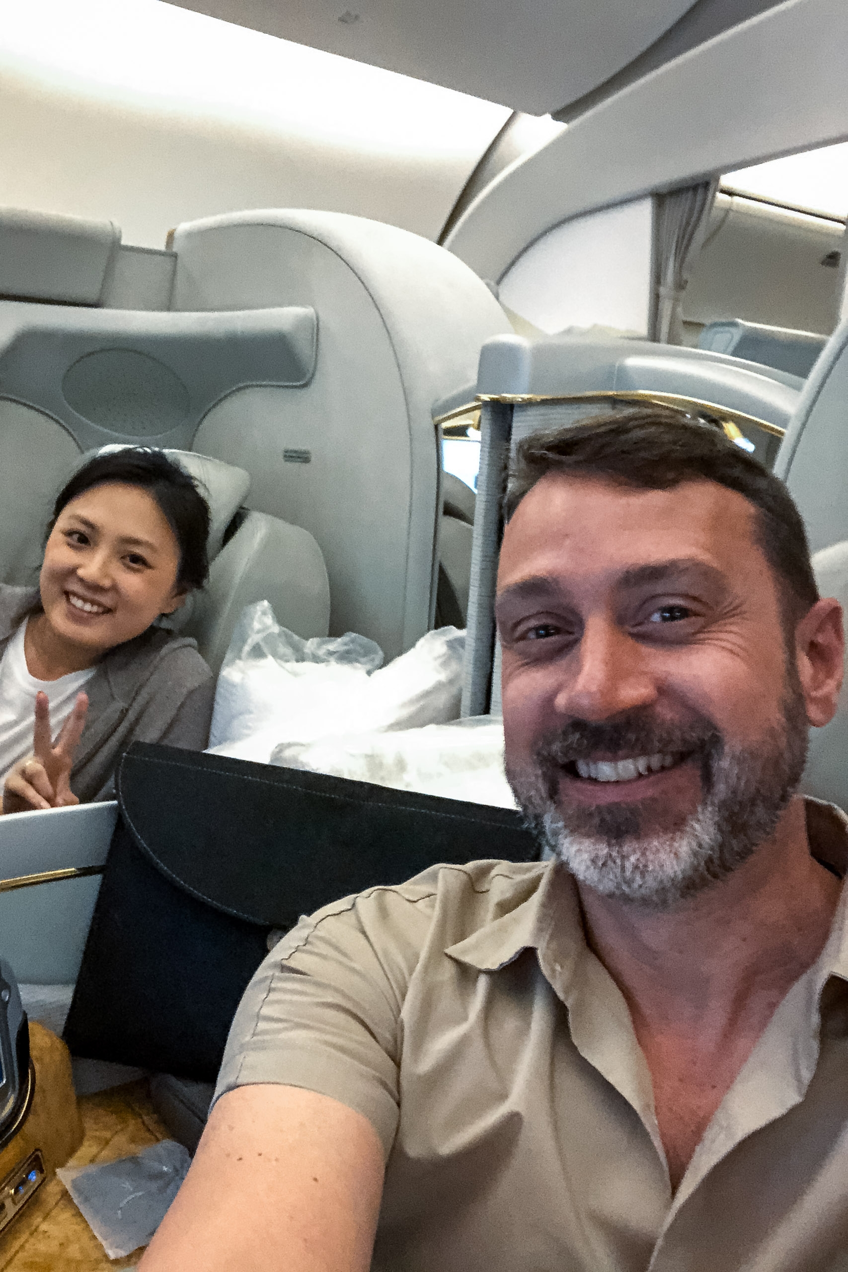 A bearded man and smiling woman take a selfie inside an airplane cabin with overhead compartments and white seats visible behind them