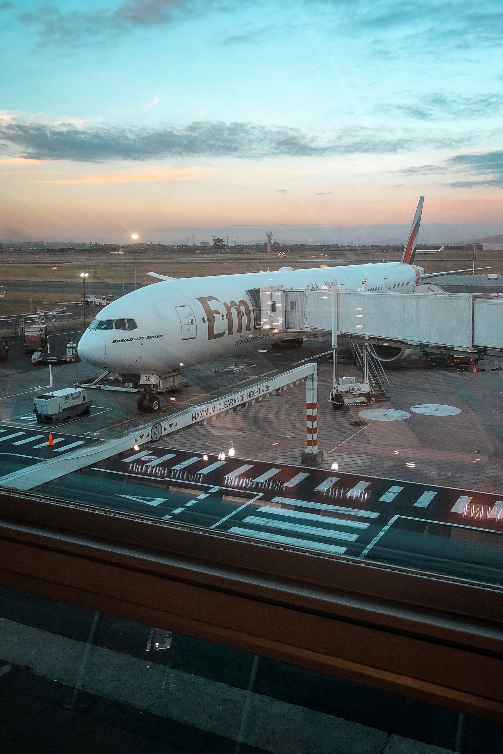 An Emirates airline aircraft is docked at a gate during twilight, with ground service vehicles and loading equipment surrounding it under a softly lit sky