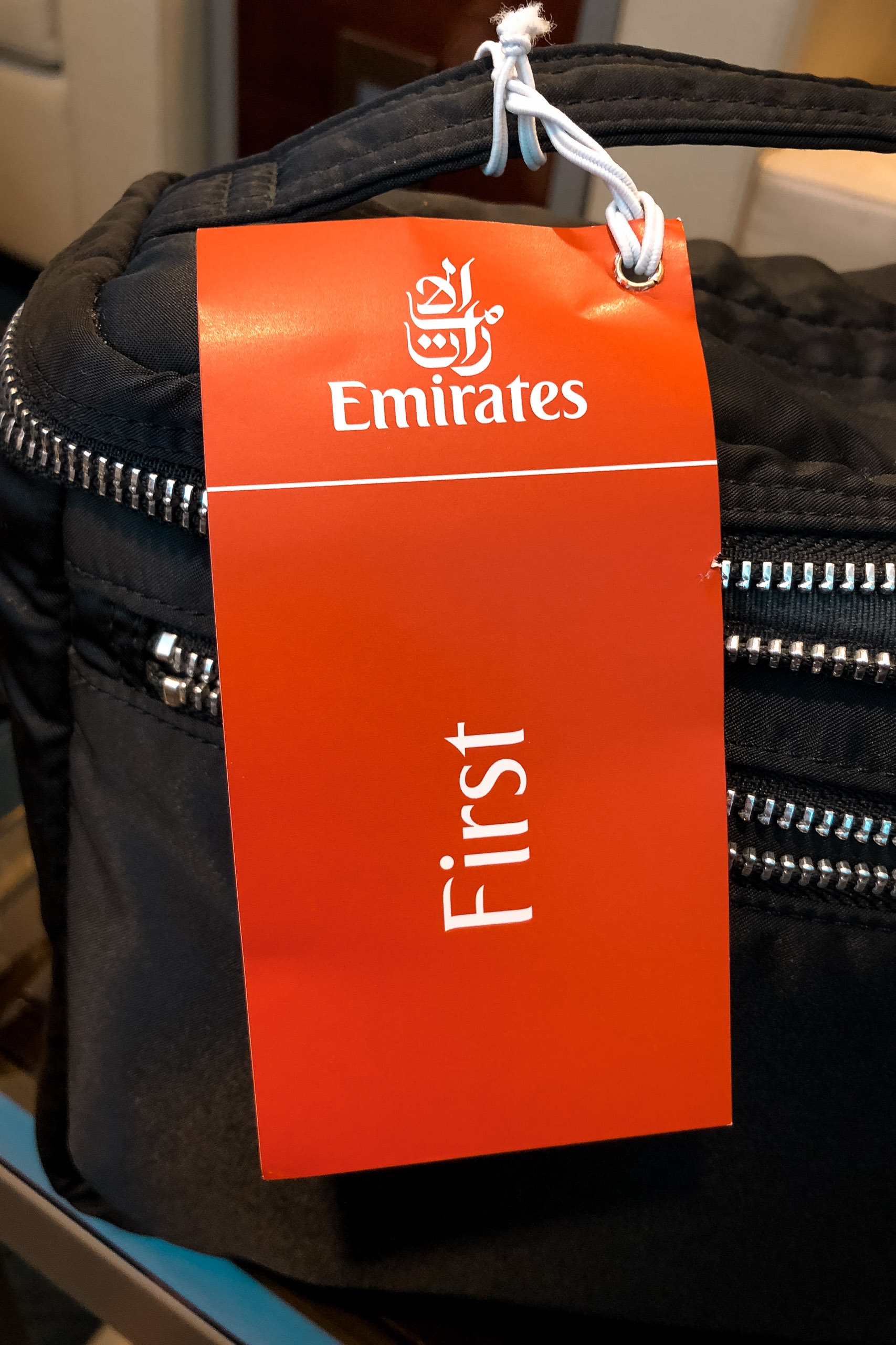 A black Emirates airline amenity bag with white zippers hangs on a plastic hanger, displaying an attached orange "Emirates First" class tag in a warm interior setting