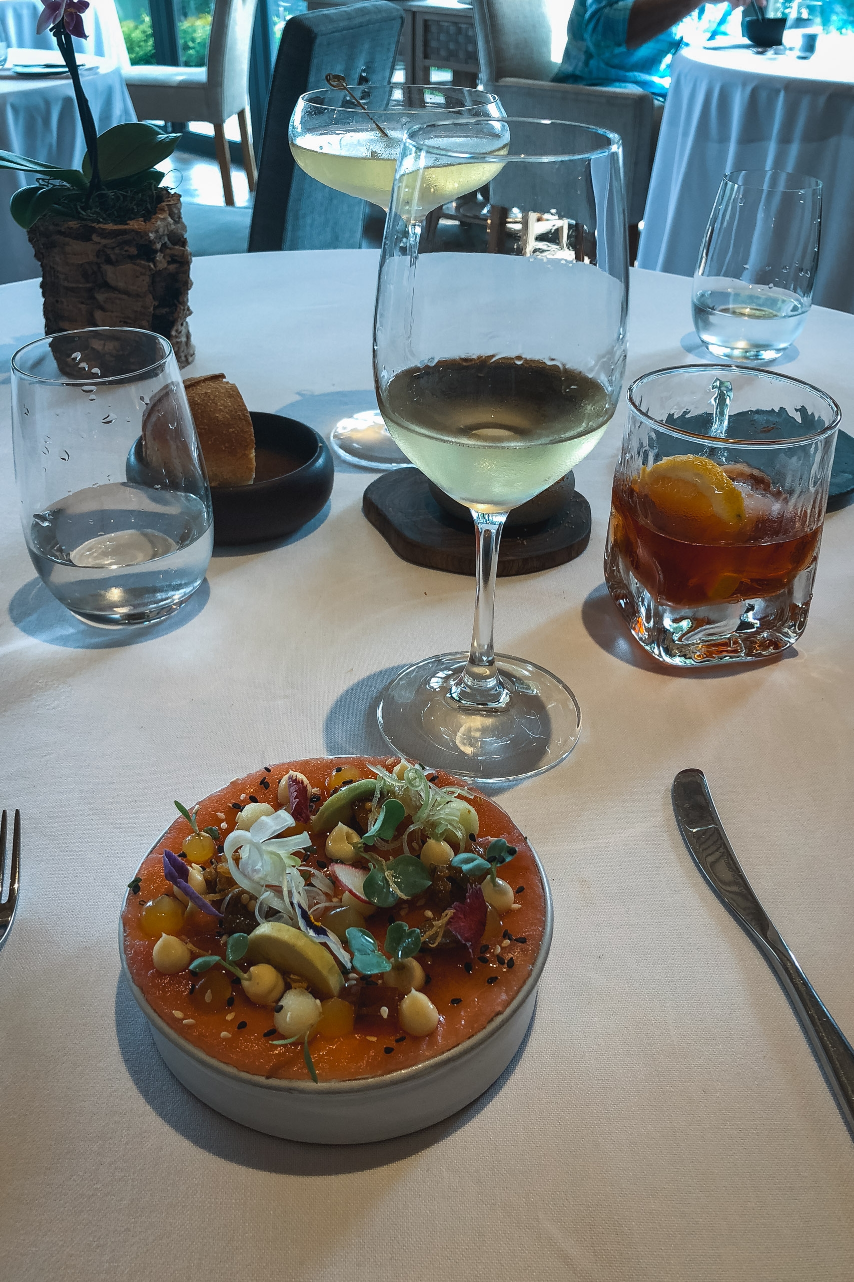A white tablecloth dining setup features a plated dish with vegetables and garnish, accompanied by glasses of white wine, water, and dark cocktail under bright natural light