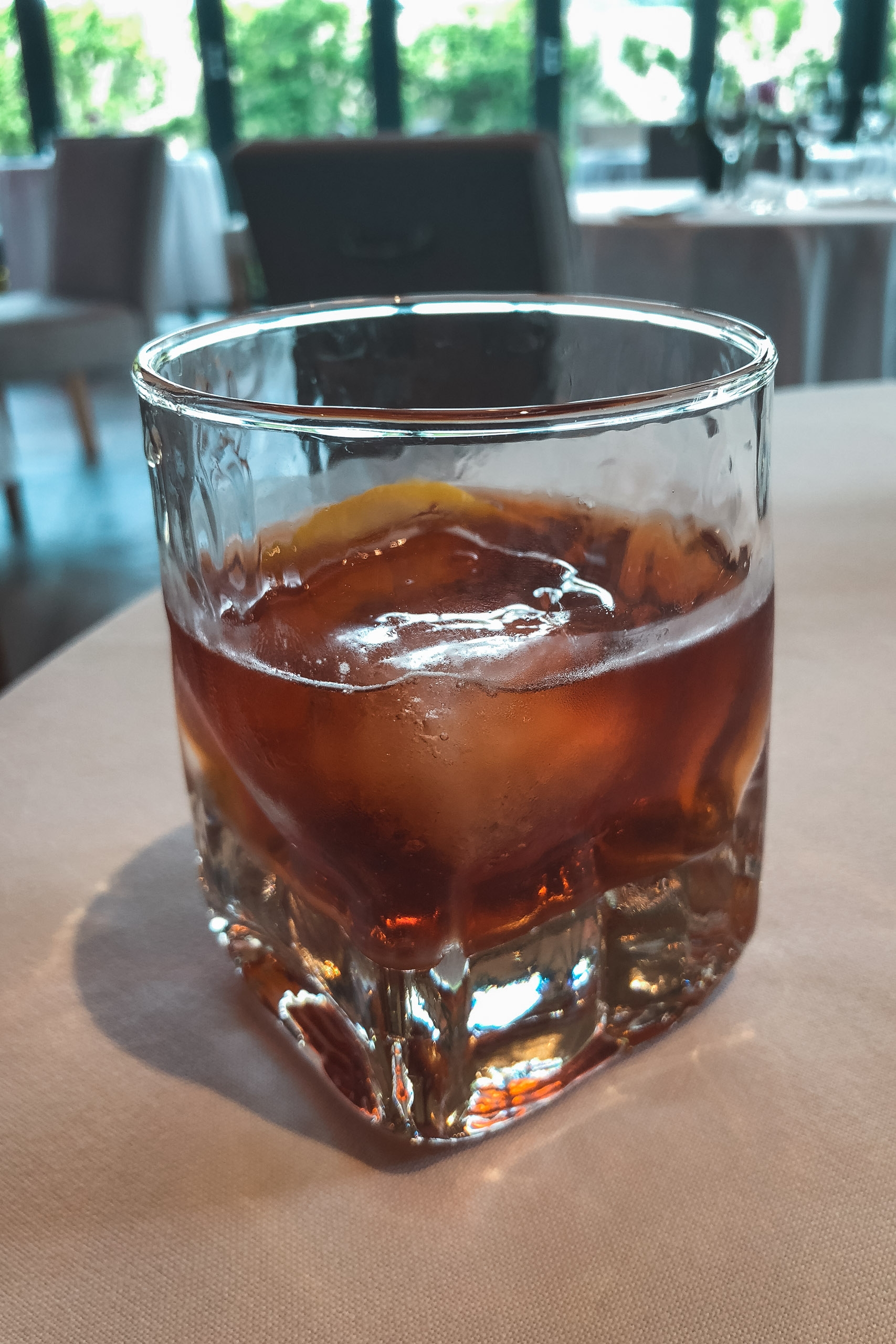 A glass of cola with ice sits on a light table in a bright, modern interior space with blurred furniture and windows in the background
