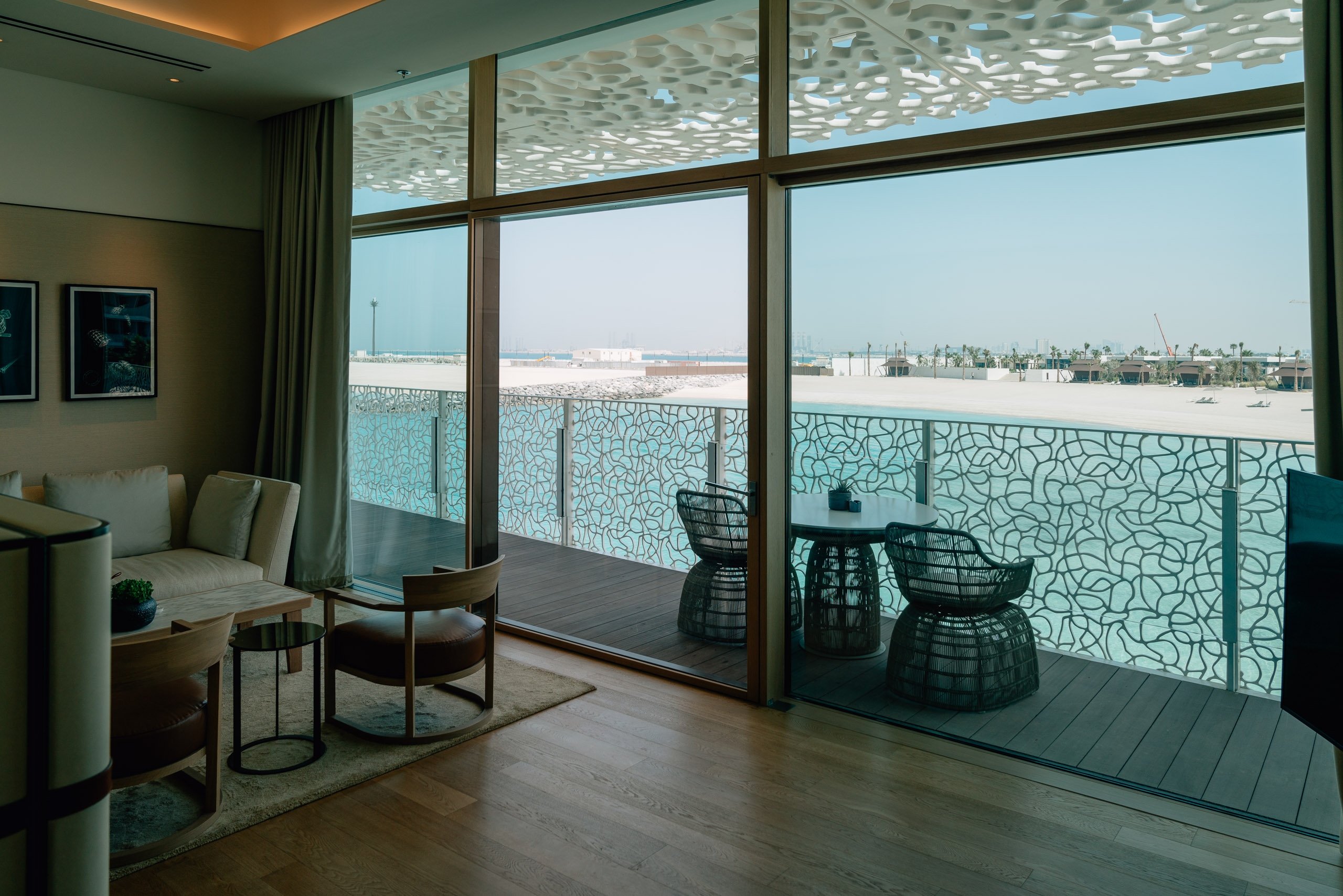 Modern luxury apartment interior with floor-to-ceiling windows overlooking a desert city landscape, featuring contemporary furniture and geometric patterned screens on a spacious balcony