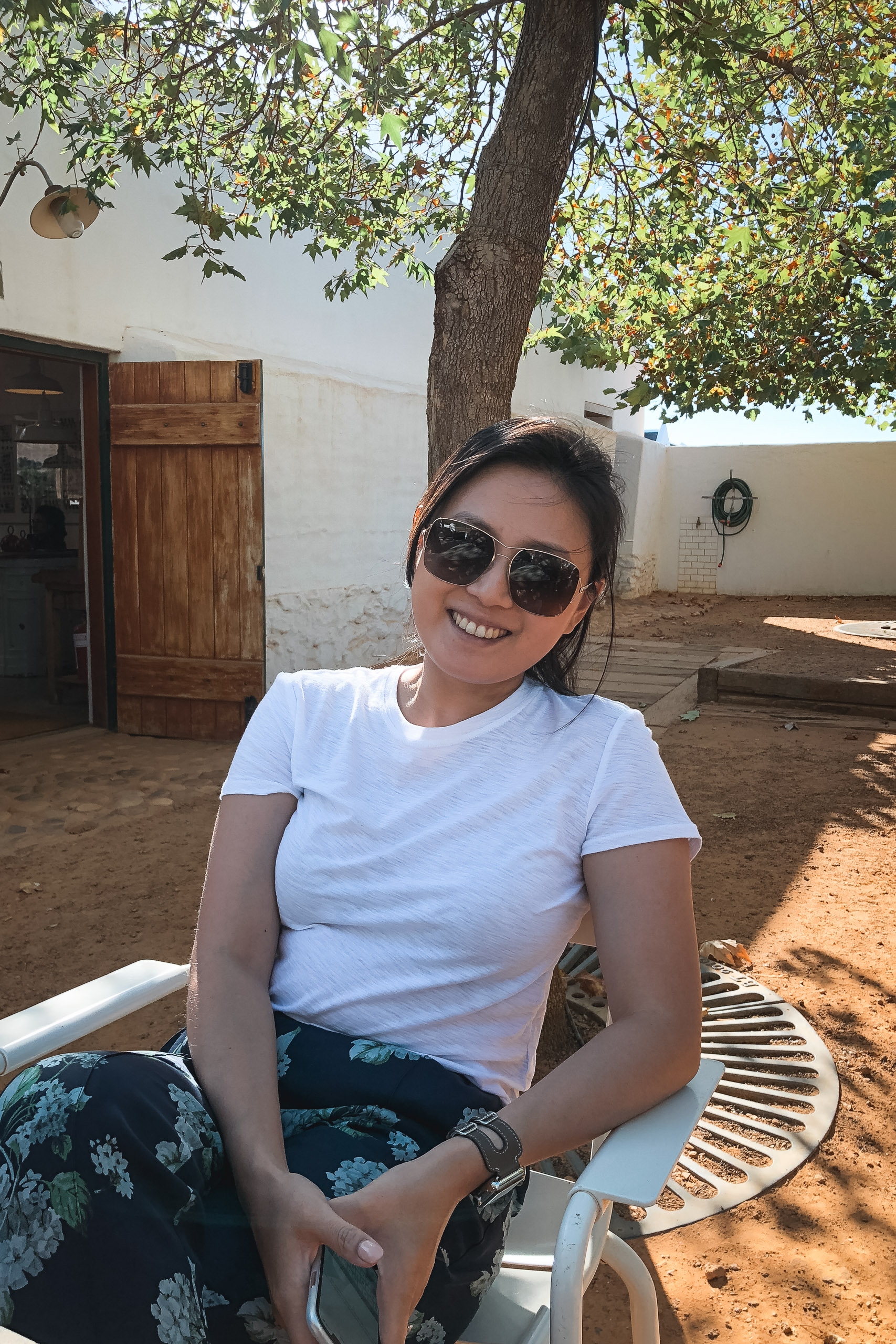 A smiling woman wearing sunglasses and a white t-shirt sits in a sandy courtyard beneath a large tree, with whitewashed buildings and wooden doors visible behind her