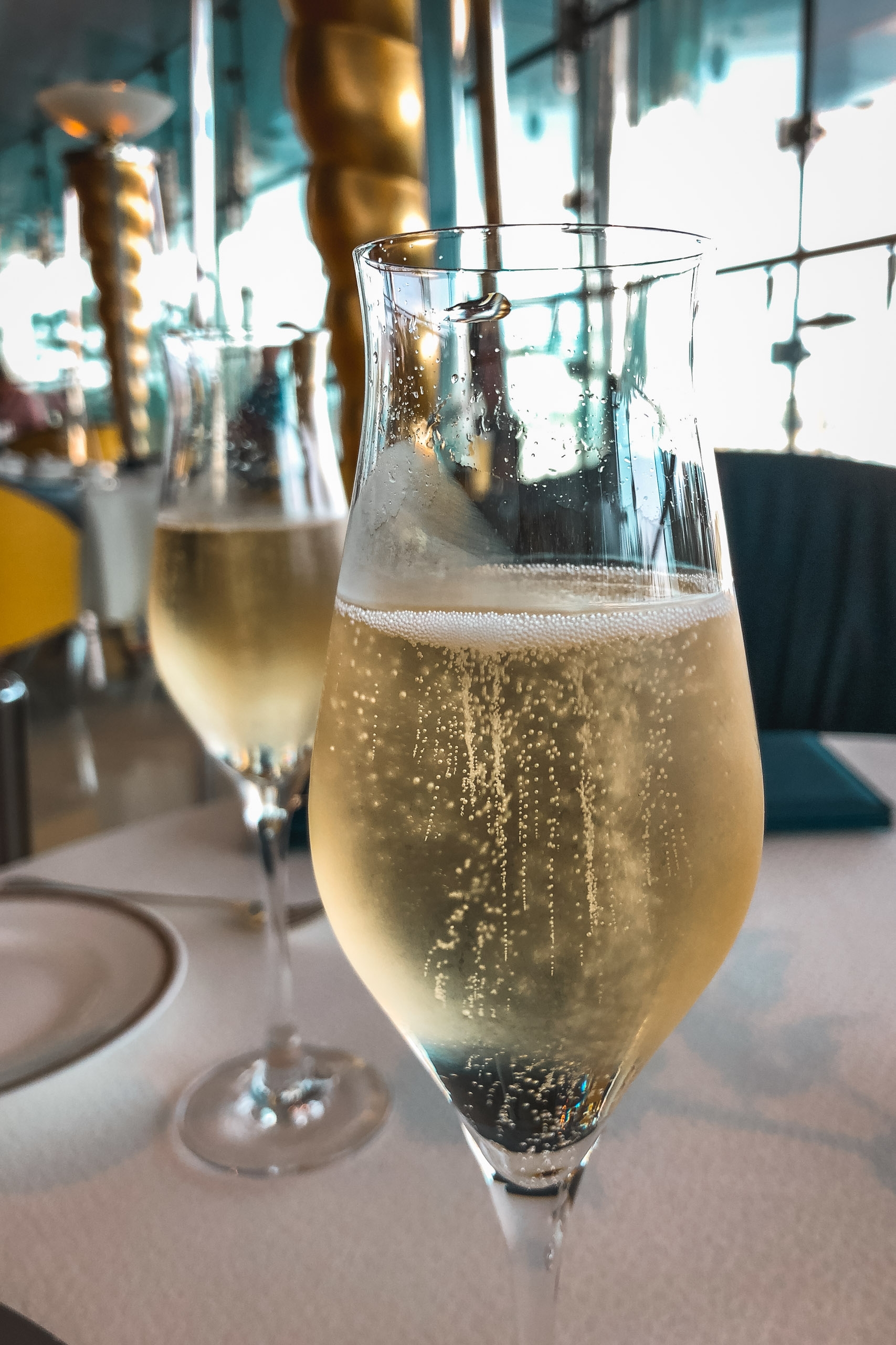 A champagne flute filled with sparkling wine sits on a white tablecloth in a bright, modern high-rise dining space with city views and blurred golden decorative elements in the background