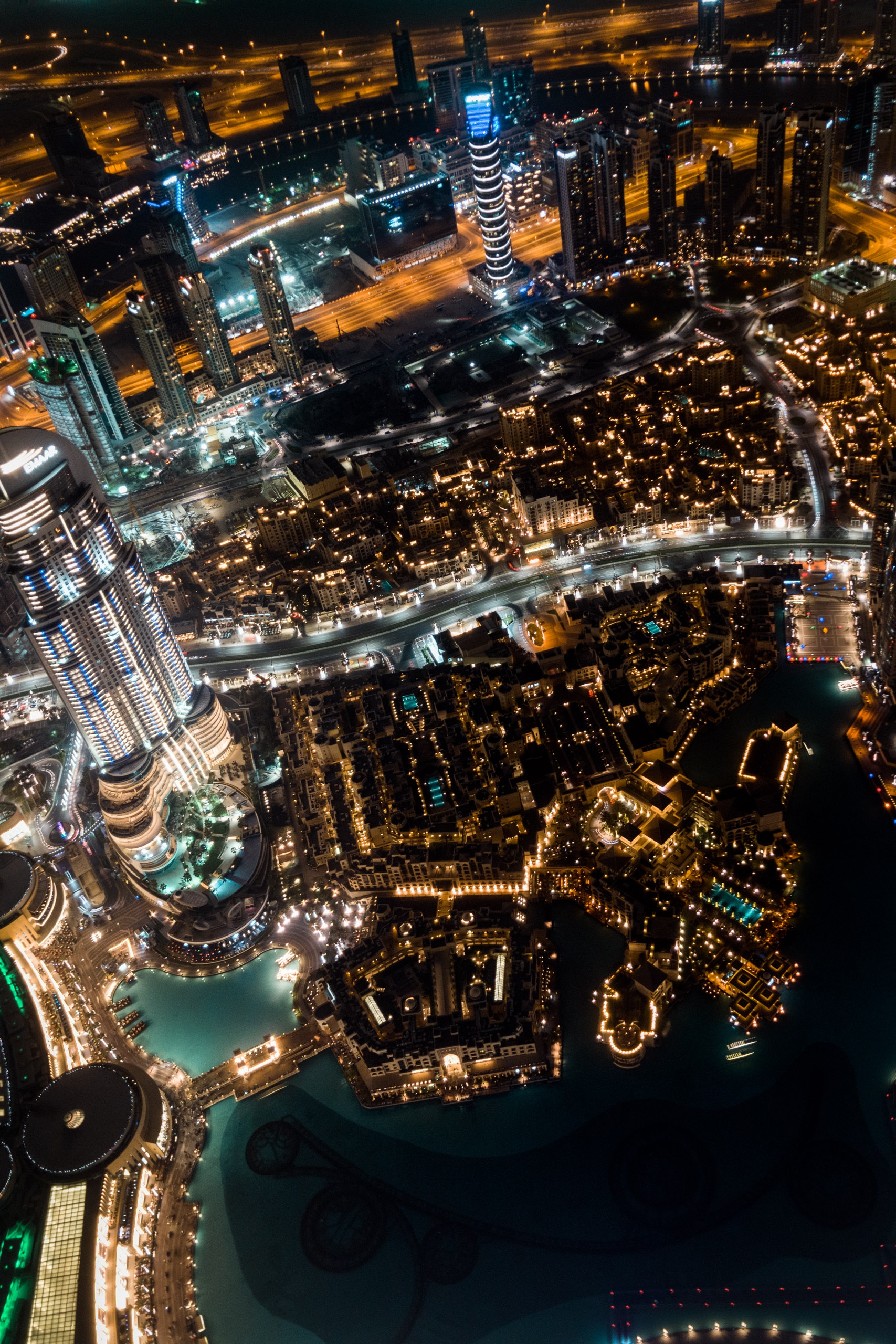 Aerial nighttime view of Dubai's illuminated skyline featuring high-rise buildings, a distinctive spiral tower, curved roadway, and turquoise water reflecting warm golden and cool blue lights below