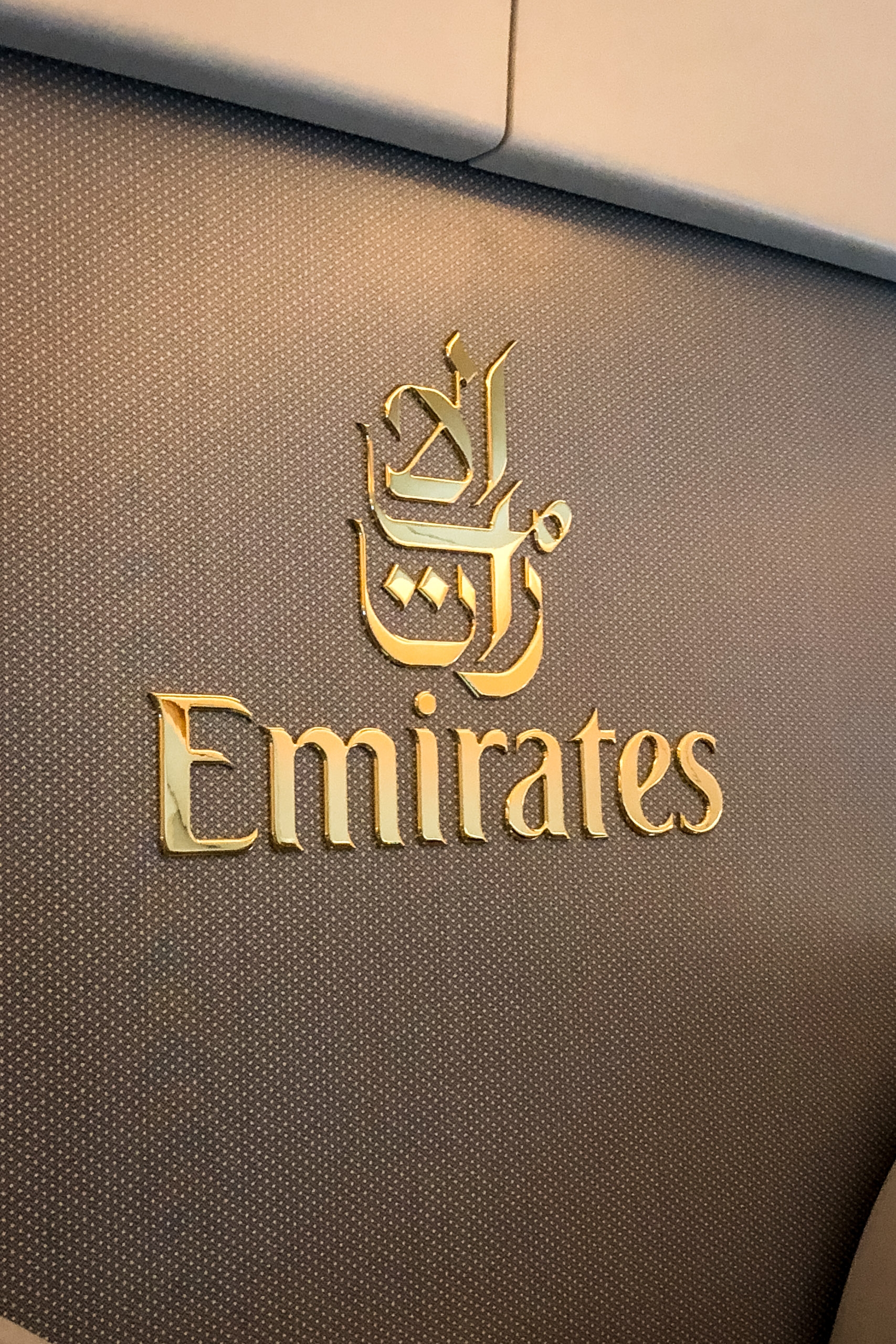 Gold Emirates logo and Arabic script embossed on textured gray upholstery, likely from an aircraft seat or interior surface with warm lighting