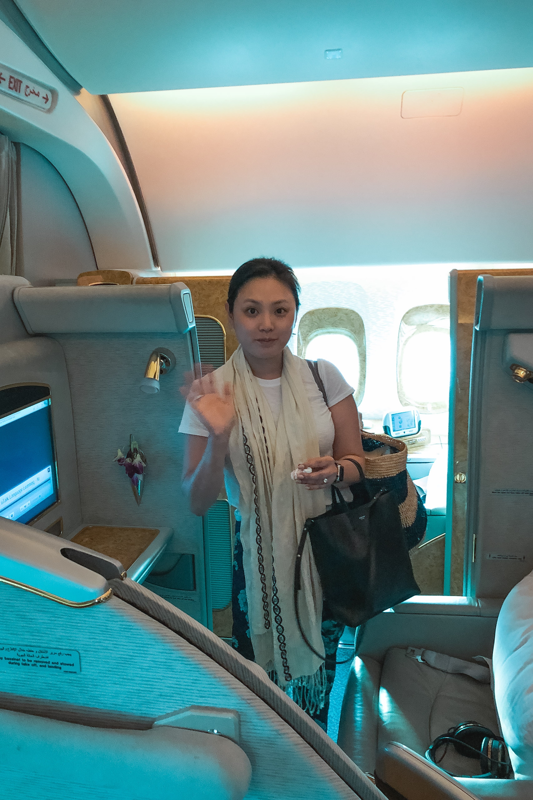 Woman standing in airplane aisle wearing light cardigan and holding black bag, bathed in turquoise cabin lighting with windows visible behind her