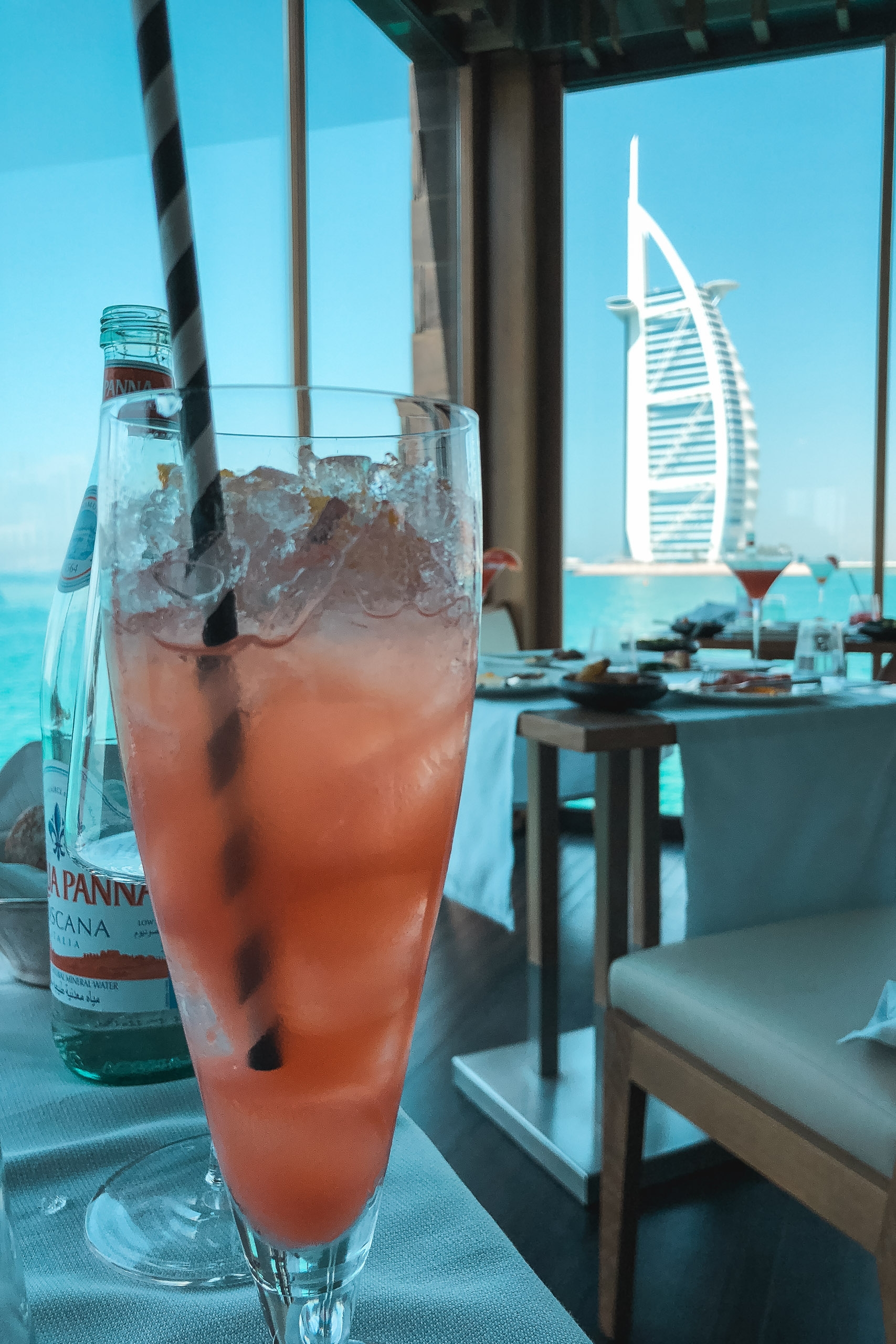 A coral-colored cocktail with ice sits on a table beside a water bottle, framing the iconic Burj Al Arab sail-shaped hotel against a clear blue sky through floor-to-ceiling windows