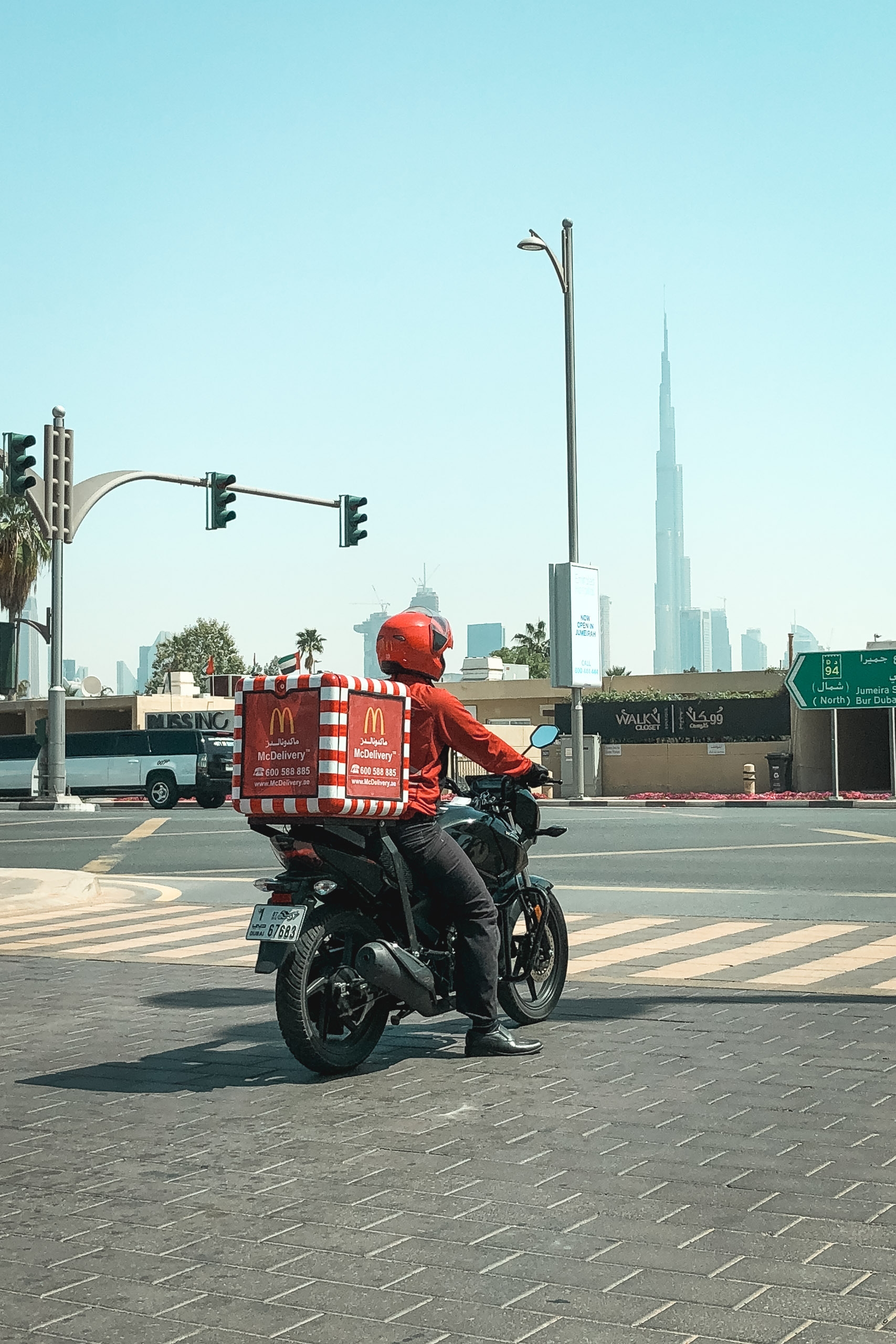 A food delivery rider in red McDonald's uniform sits on a motorcycle at a Dubai street intersection, with the Burj Khalifa towering in the hazy background skyline