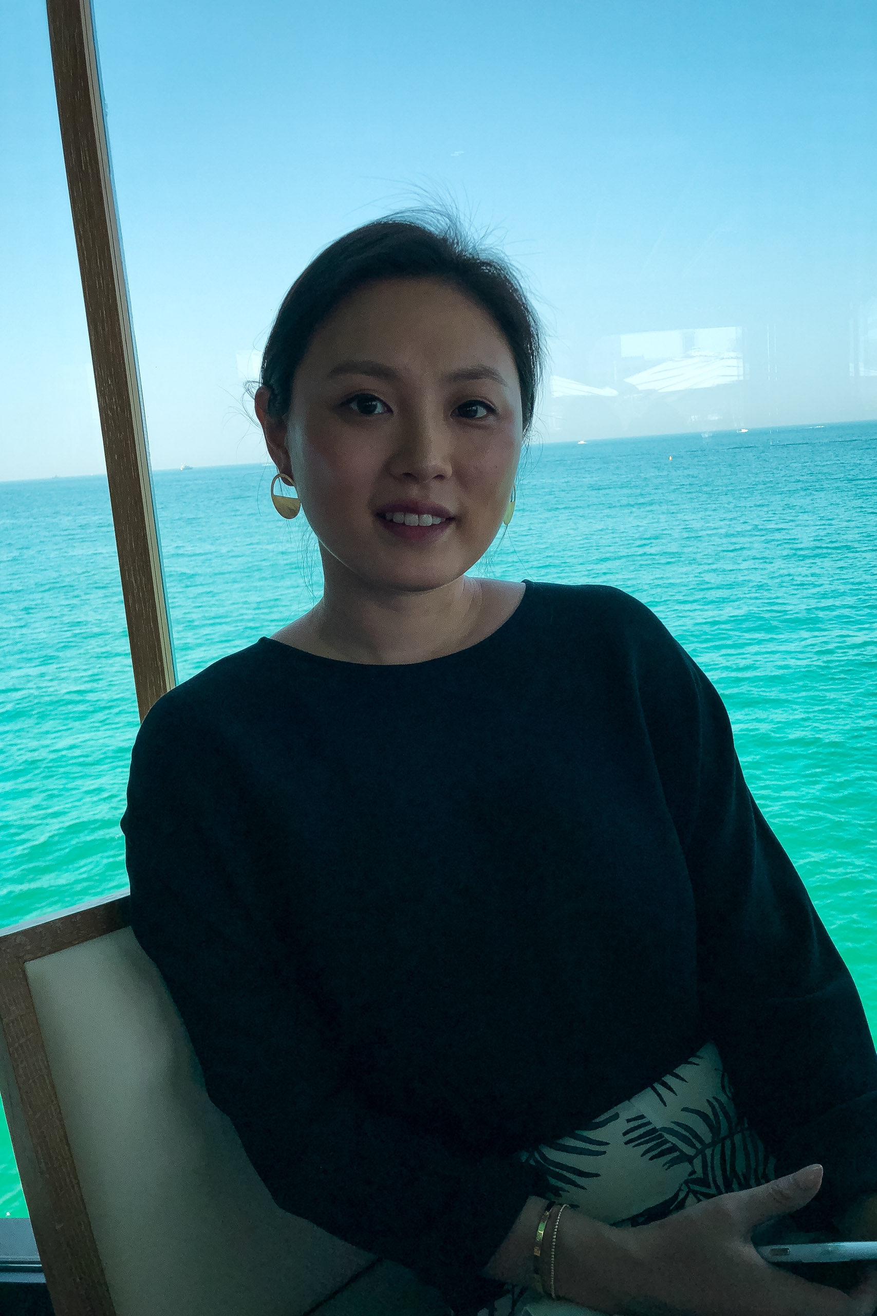 Woman in a black long-sleeve shirt smiles at the camera while seated on a boat, with turquoise ocean water and clear sky behind her