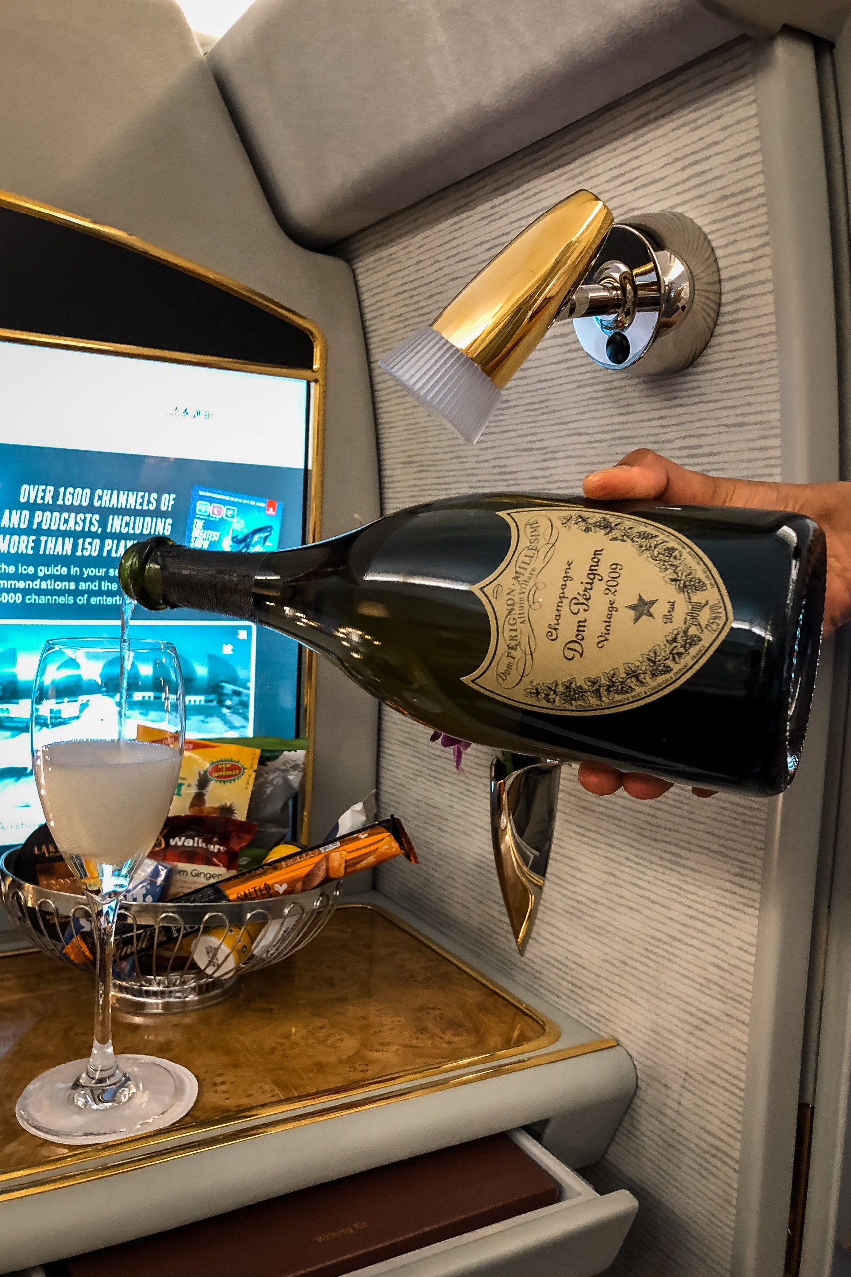 A luxury aircraft cabin features a champagne bottle held against a textured wall panel with a golden spotlight above, beside a cocktail and snacks on a wooden tray table with a glowing screen visible in the background
