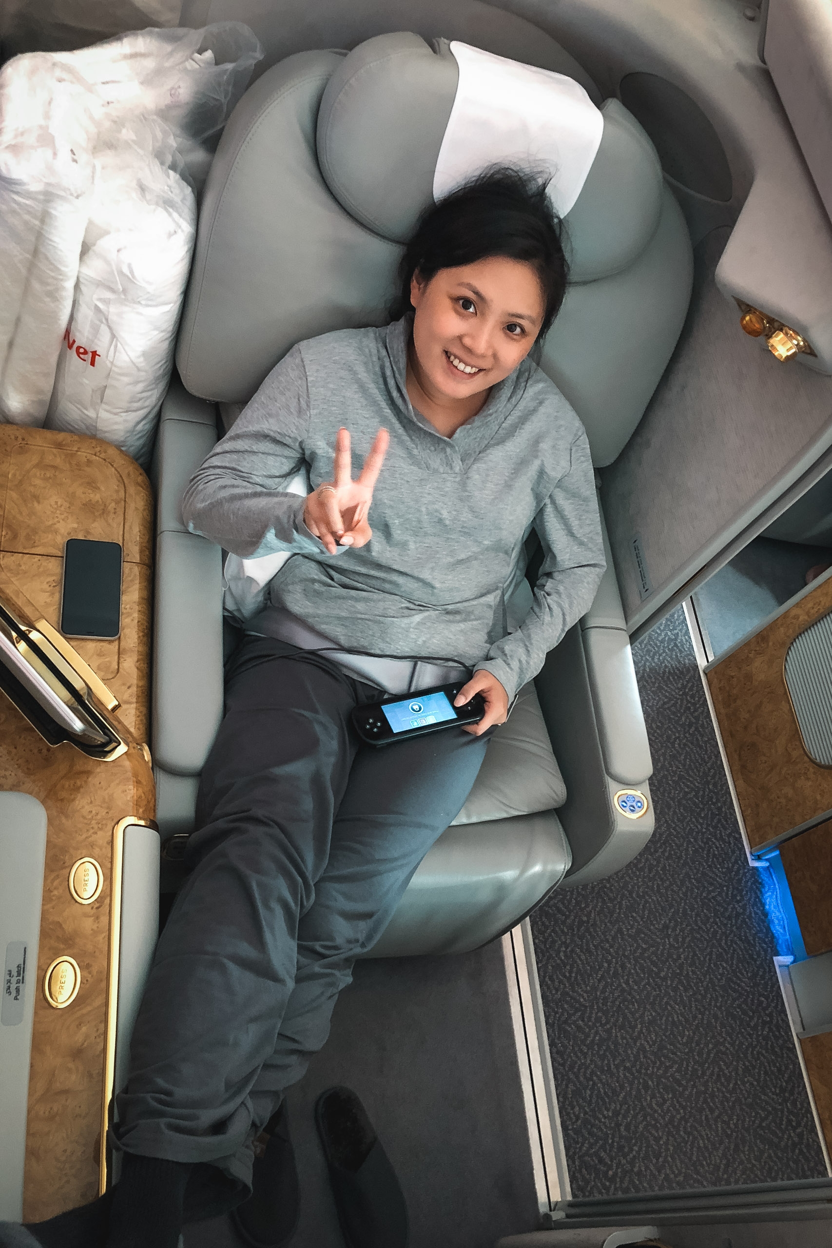 A woman reclines in a gray airplane seat wearing a gray hoodie, making a peace sign while holding a mobile device, photographed from directly above