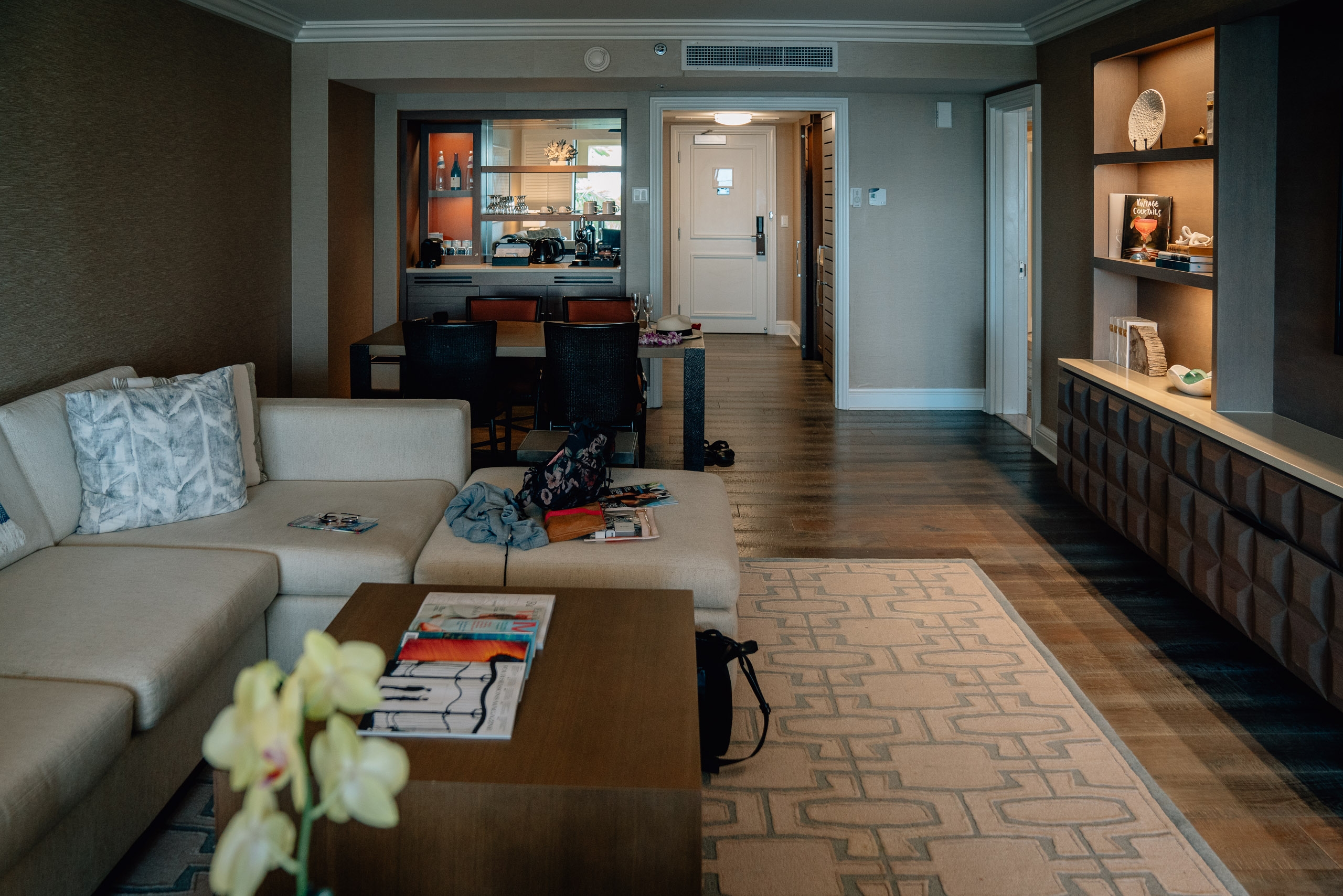 A spacious hotel suite interior featuring a beige sectional sofa, wooden coffee table with magazines, warm wood flooring, and illuminated display shelves on the right wall, with a kitchen and entryway visible in the background