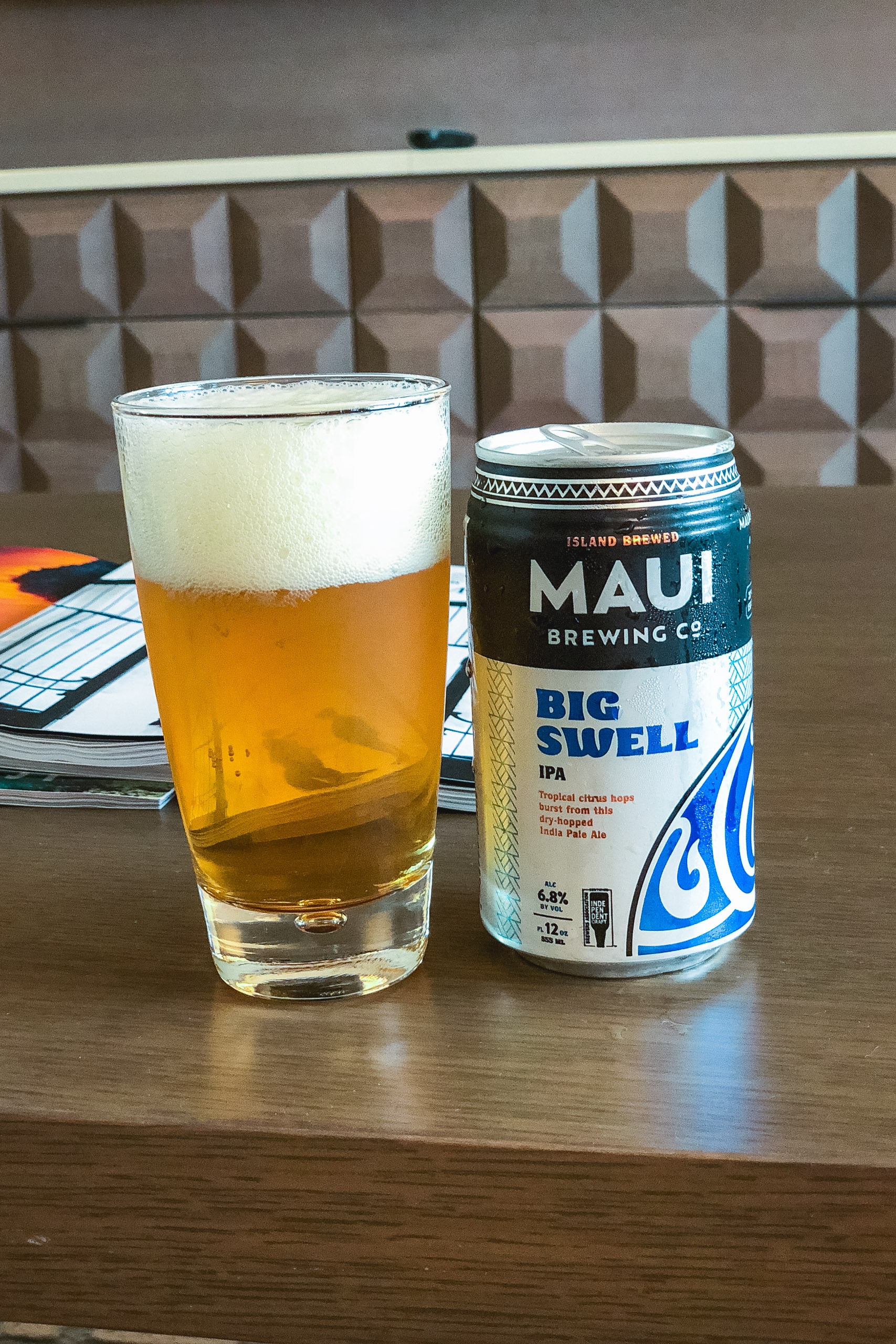 A pint glass filled with golden IPA beer and foam sits beside a Maui Brewing Company Big Swell can on a wooden table, with a geometric wall panel and notebook visible in the background.