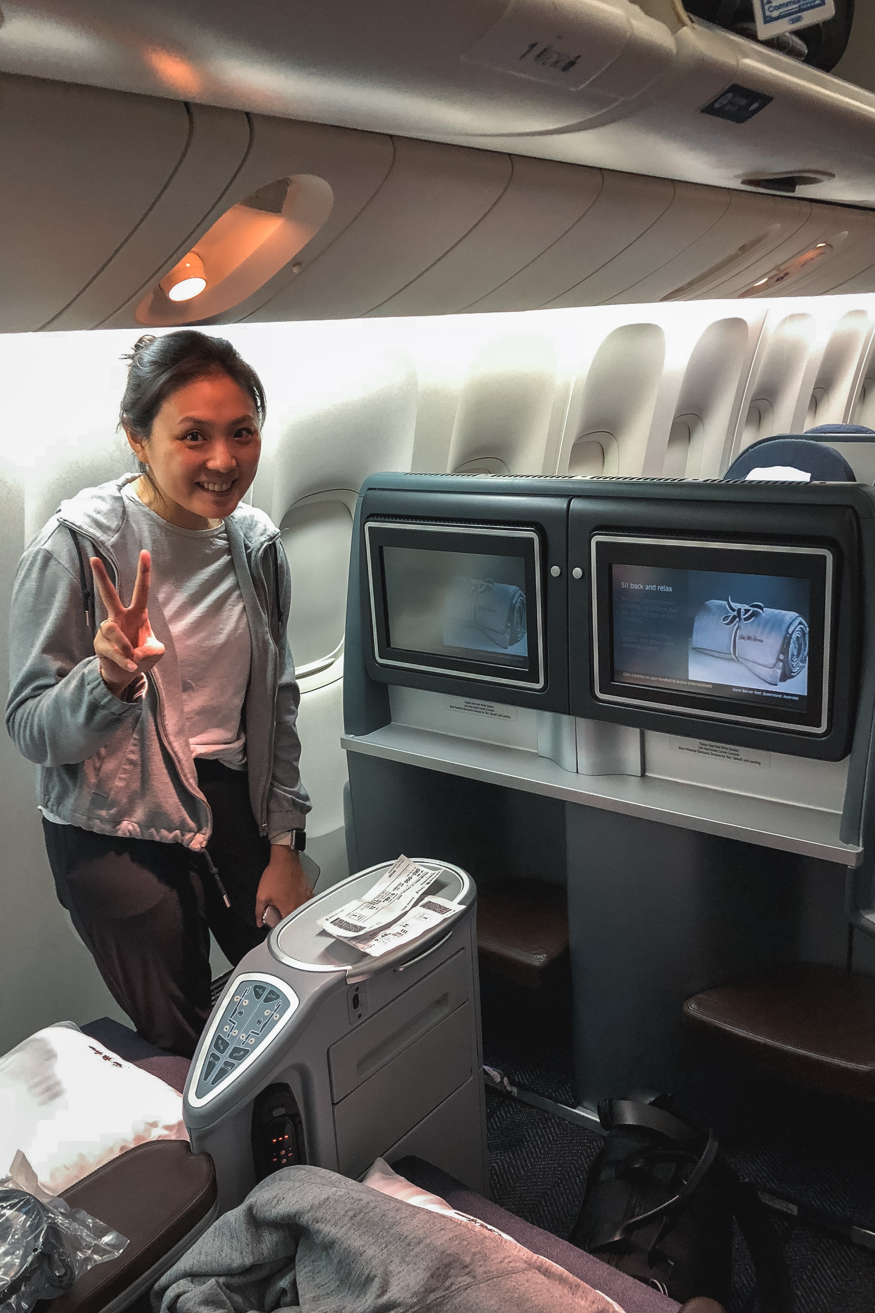 Woman gestures peacefully inside an airplane cabin next to dual entertainment screens and overhead luggage compartments with warm cabin lighting