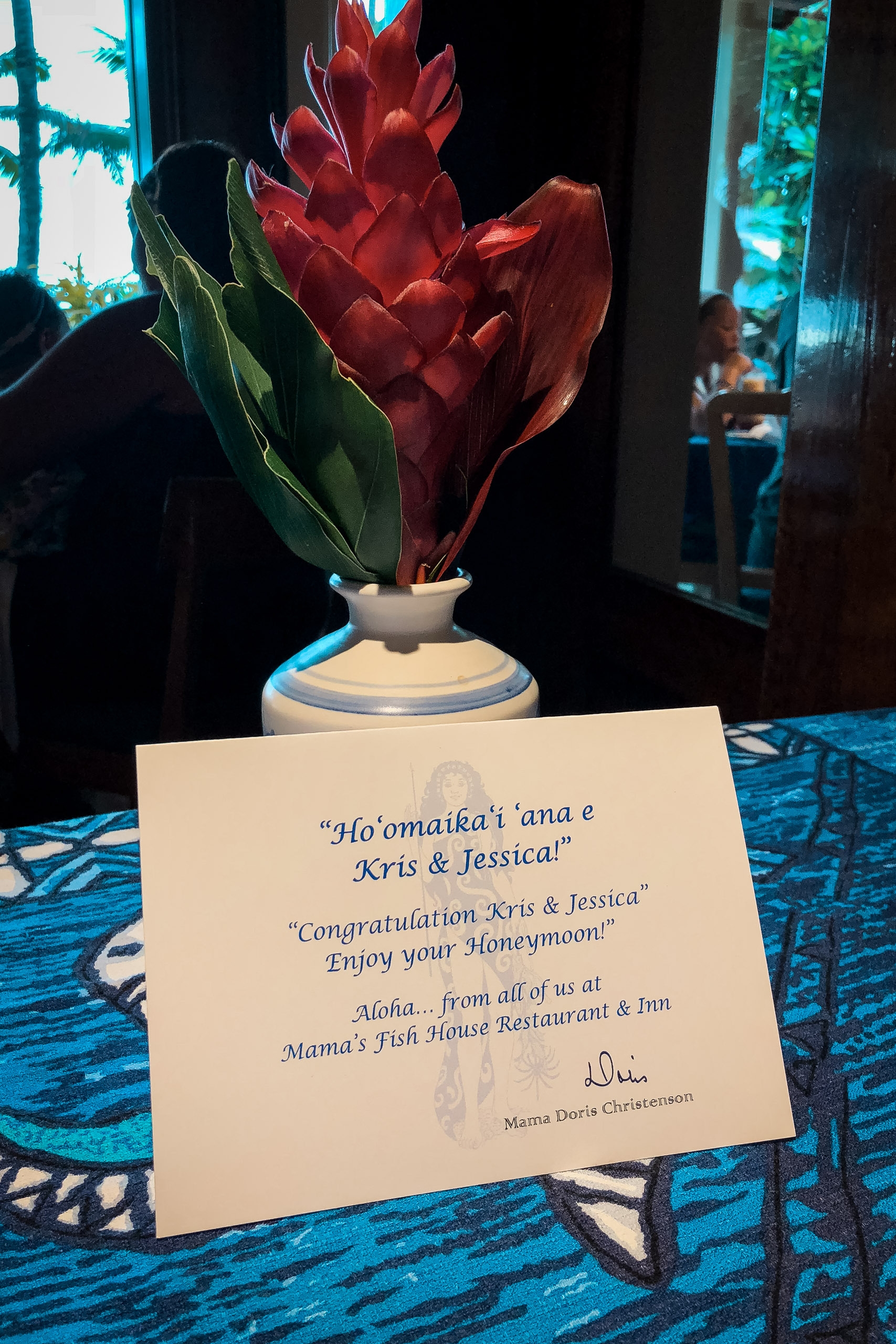 A vibrant red ginger flower arrangement in a white ceramic vase sits beside a congratulatory card on a blue patterned tablecloth at an indoor event, with turquoise-lit windows visible in the background.