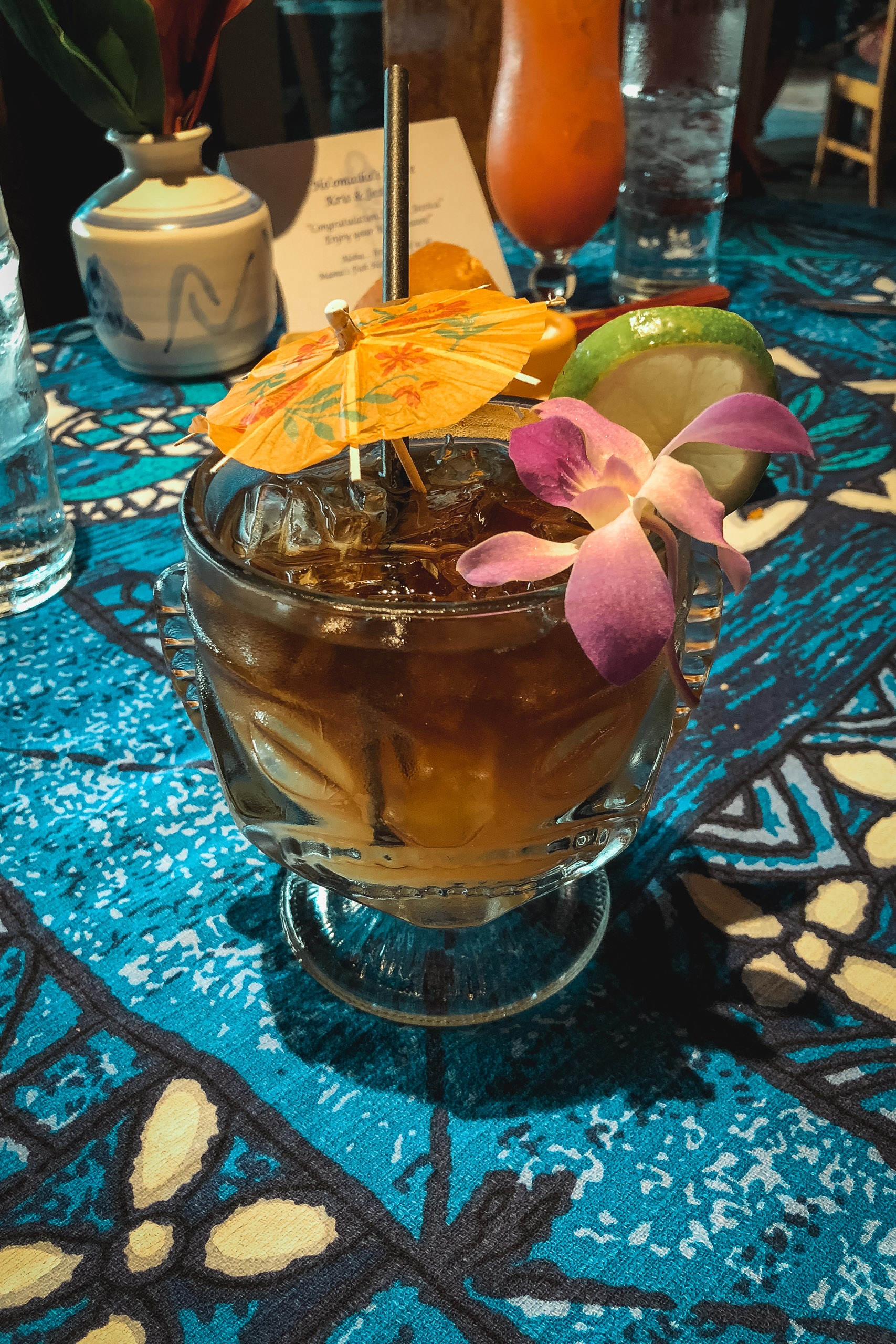 A tropical cocktail garnished with orange slices, lime, and pink orchid petals sits on a vibrant blue patterned tablecloth at an upscale restaurant or resort setting
