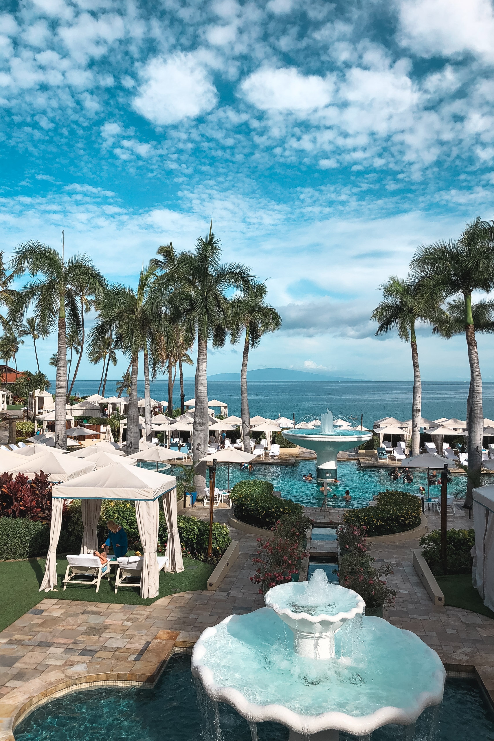 Oceanfront resort pool area with tall palm trees, white umbrellas, and turquoise water overlooking a calm sea under partly cloudy skies
