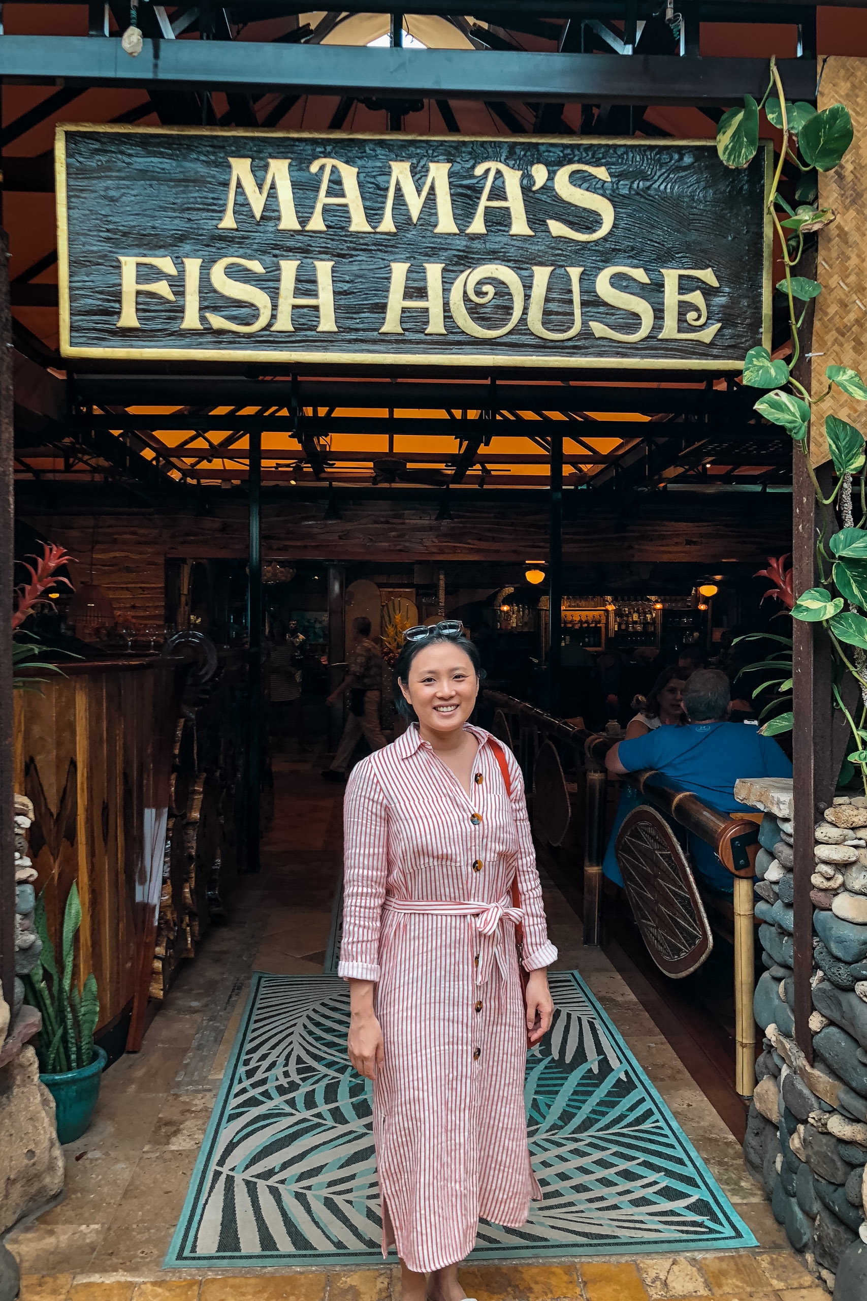 A woman in a pink striped shirt stands smiling at the entrance of Mama's Fish House restaurant, framed by a wooden storefront with tropical plants and a geometric patterned mat below