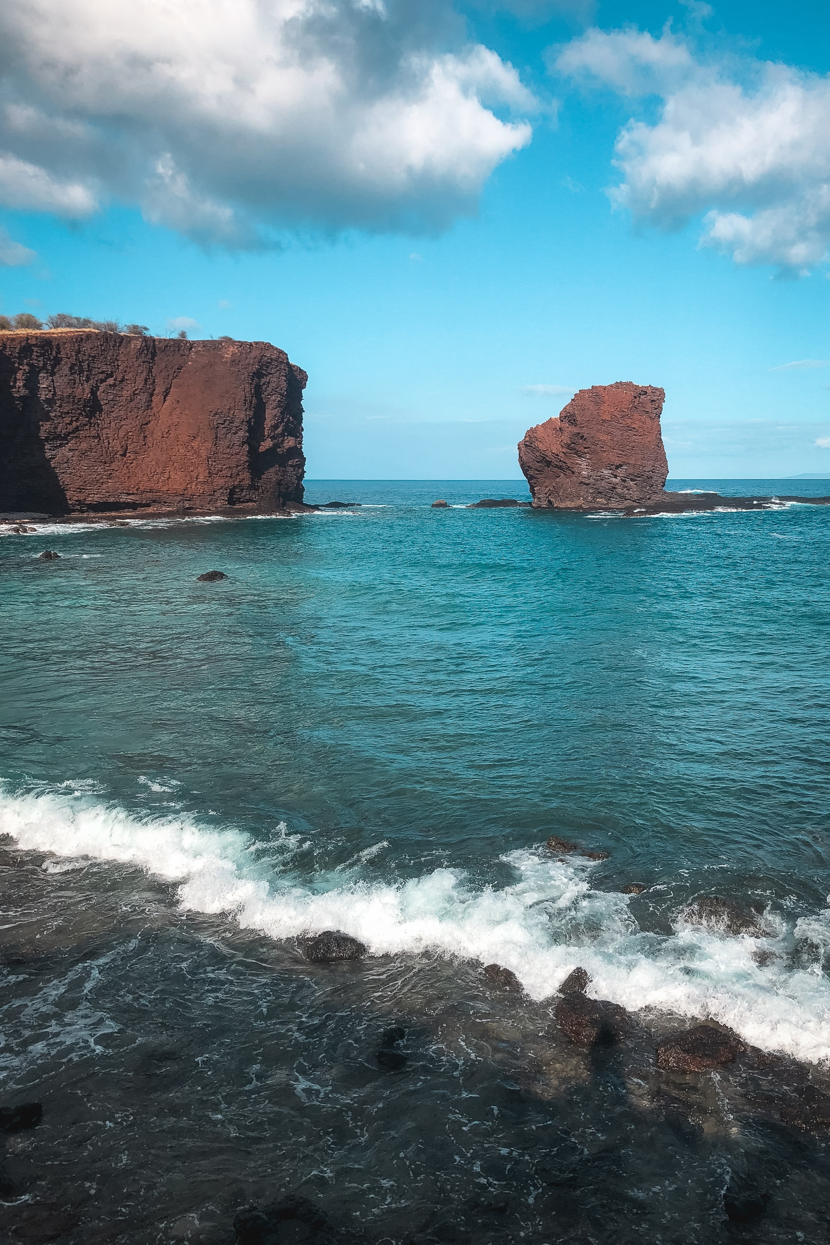 Turquoise ocean waves roll onto a rocky shore beneath two distinctive red volcanic rock formations rising from the clear blue water under a partly cloudy sky