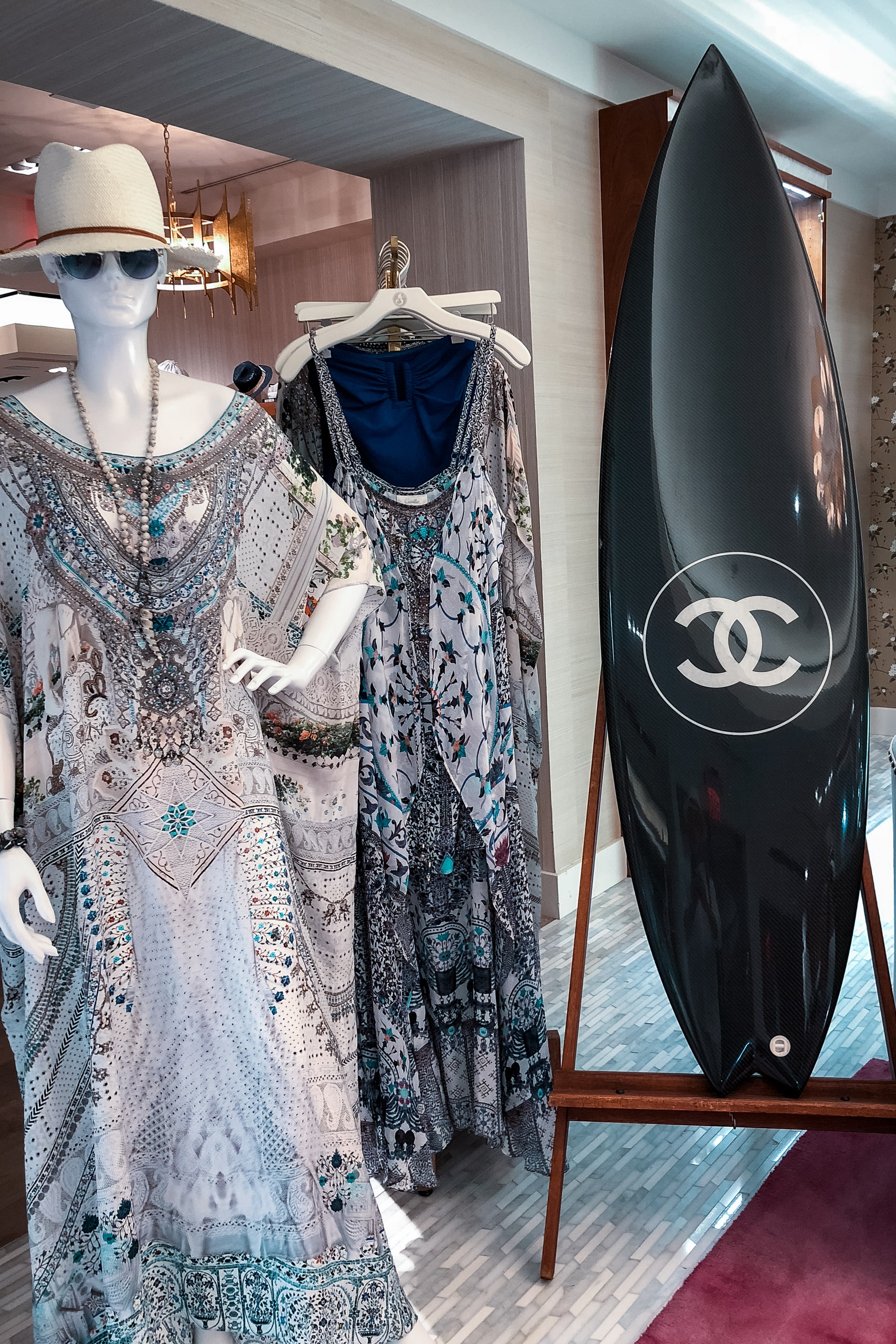 A luxury boutique display features mannequins wearing ornate patterned garments and accessories alongside a black surfboard emblazoned with the Chanel logo, merging high fashion with island lifestyle aesthetics