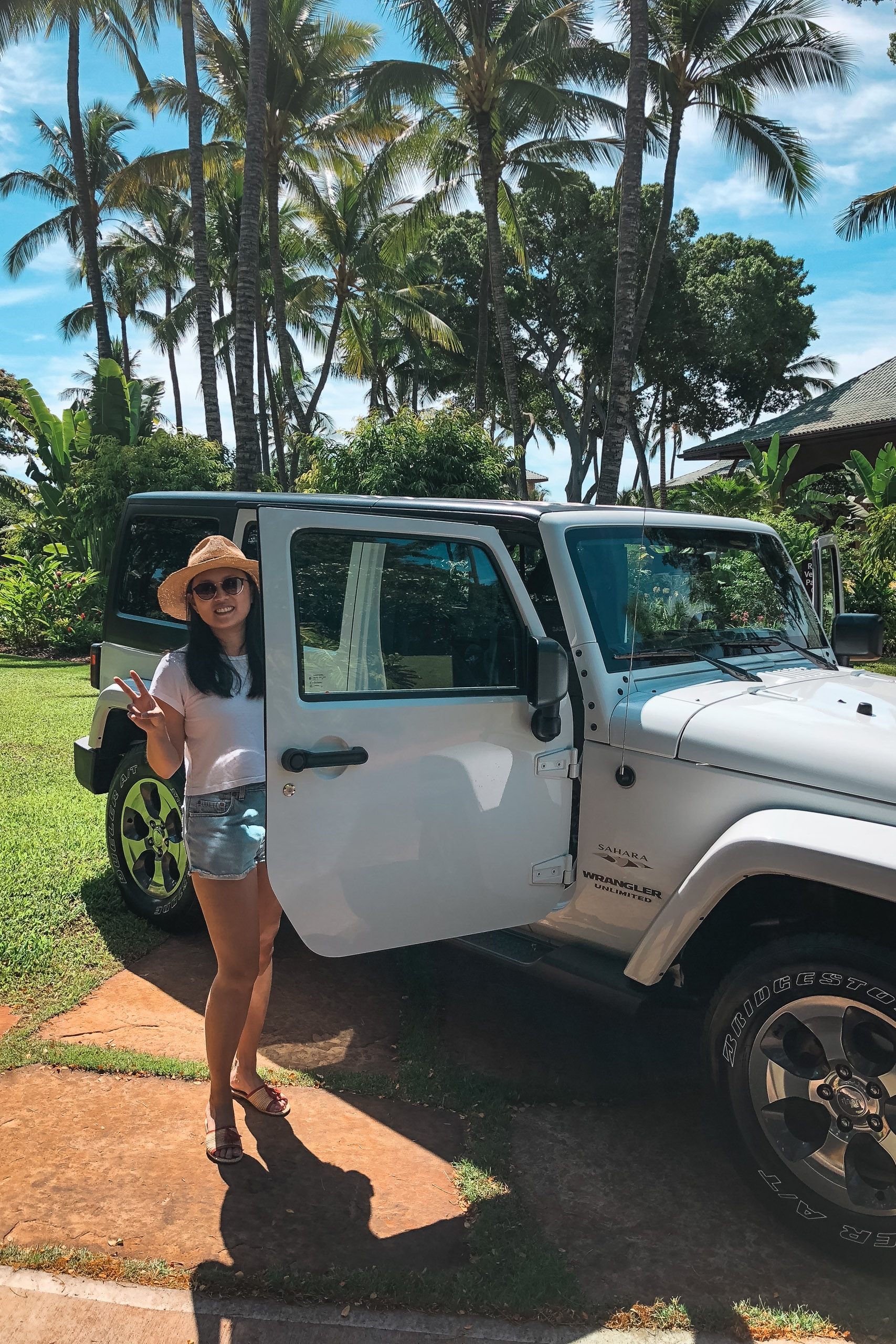 A woman in a straw hat and pink top stands beside a white Jeep in a tropical driveway lined with tall palm trees under bright blue sky