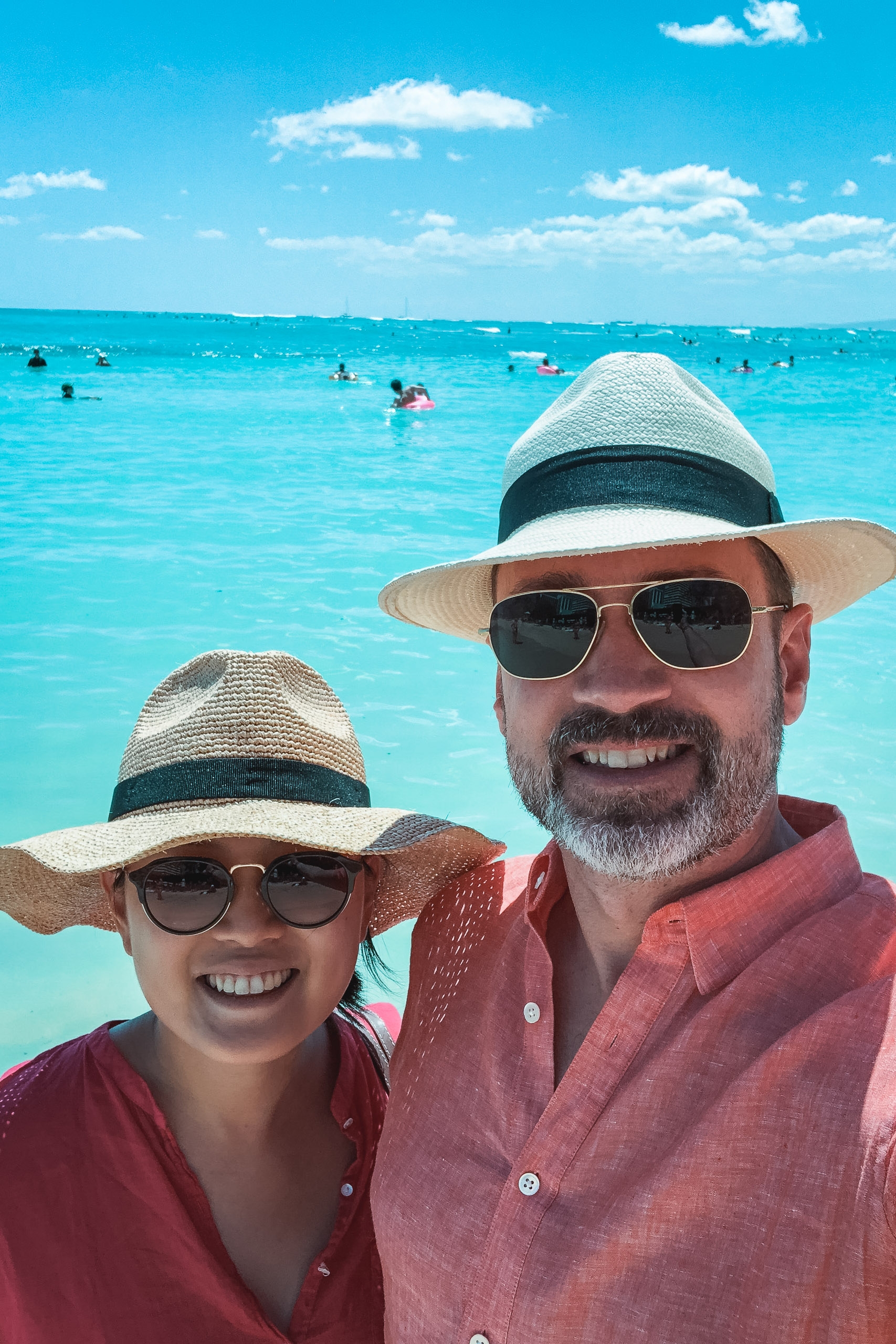 Two people wearing straw hats and sunglasses smile at the camera with turquoise ocean and swimmers visible behind them under a bright blue sky