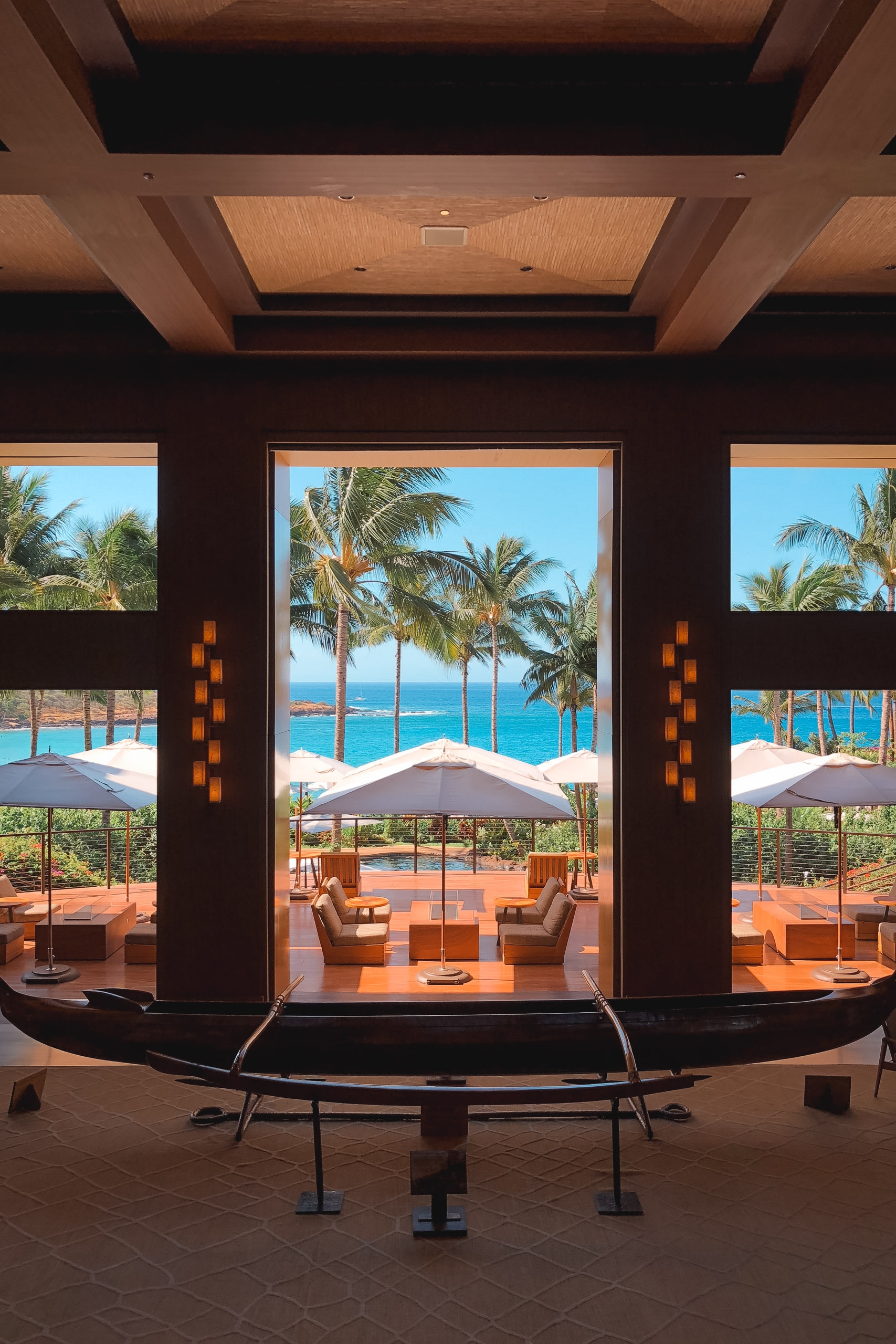 A luxurious resort interior frames a stunning coastal view through floor-to-ceiling windows, with palm trees, umbrellas, and turquoise ocean visible beyond the modern architectural space