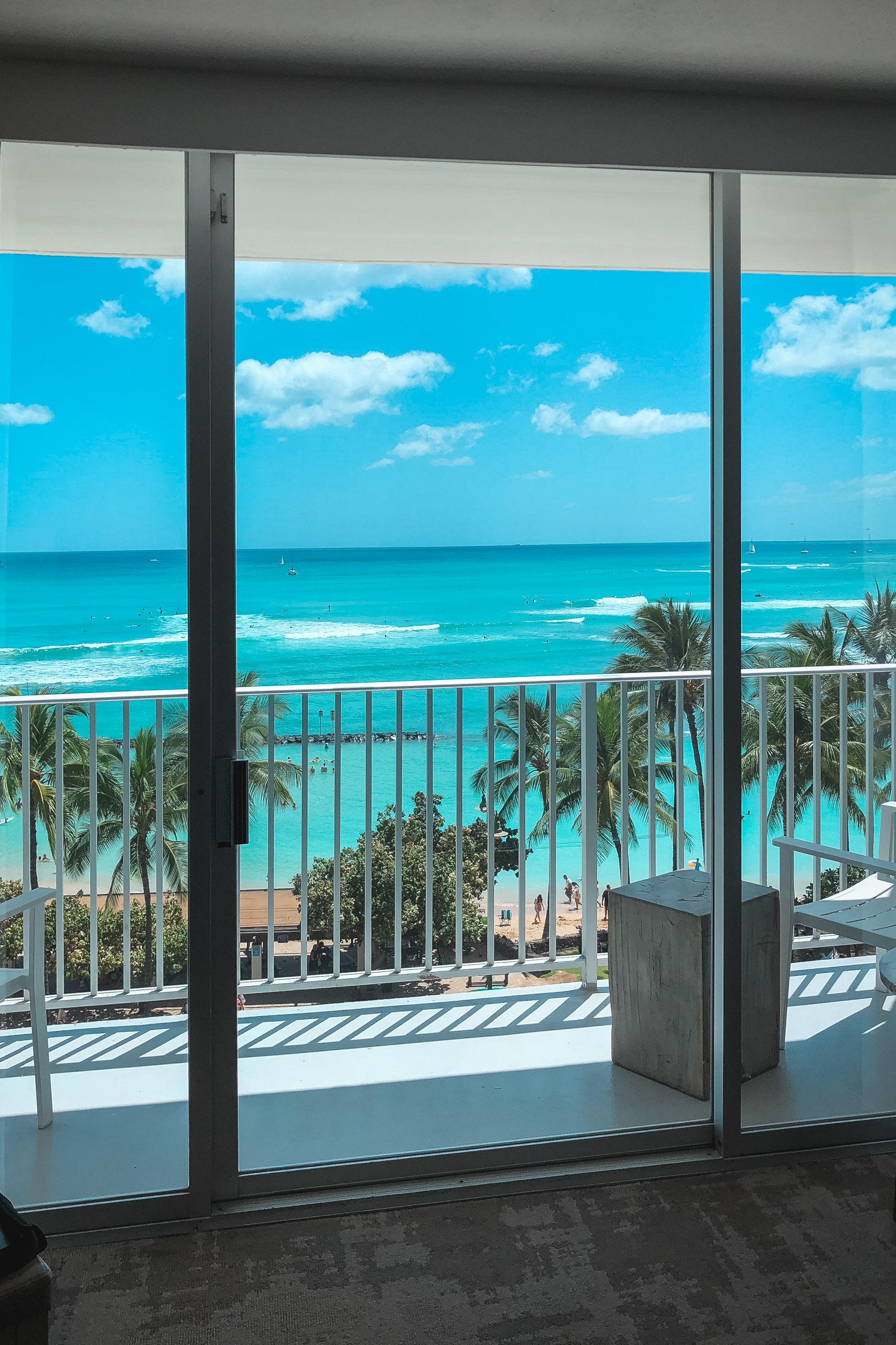 Floor-to-ceiling windows frame a tropical coastal view with turquoise waters, white-sand beach, palm trees, and puffy clouds under bright daylight from a high-rise balcony