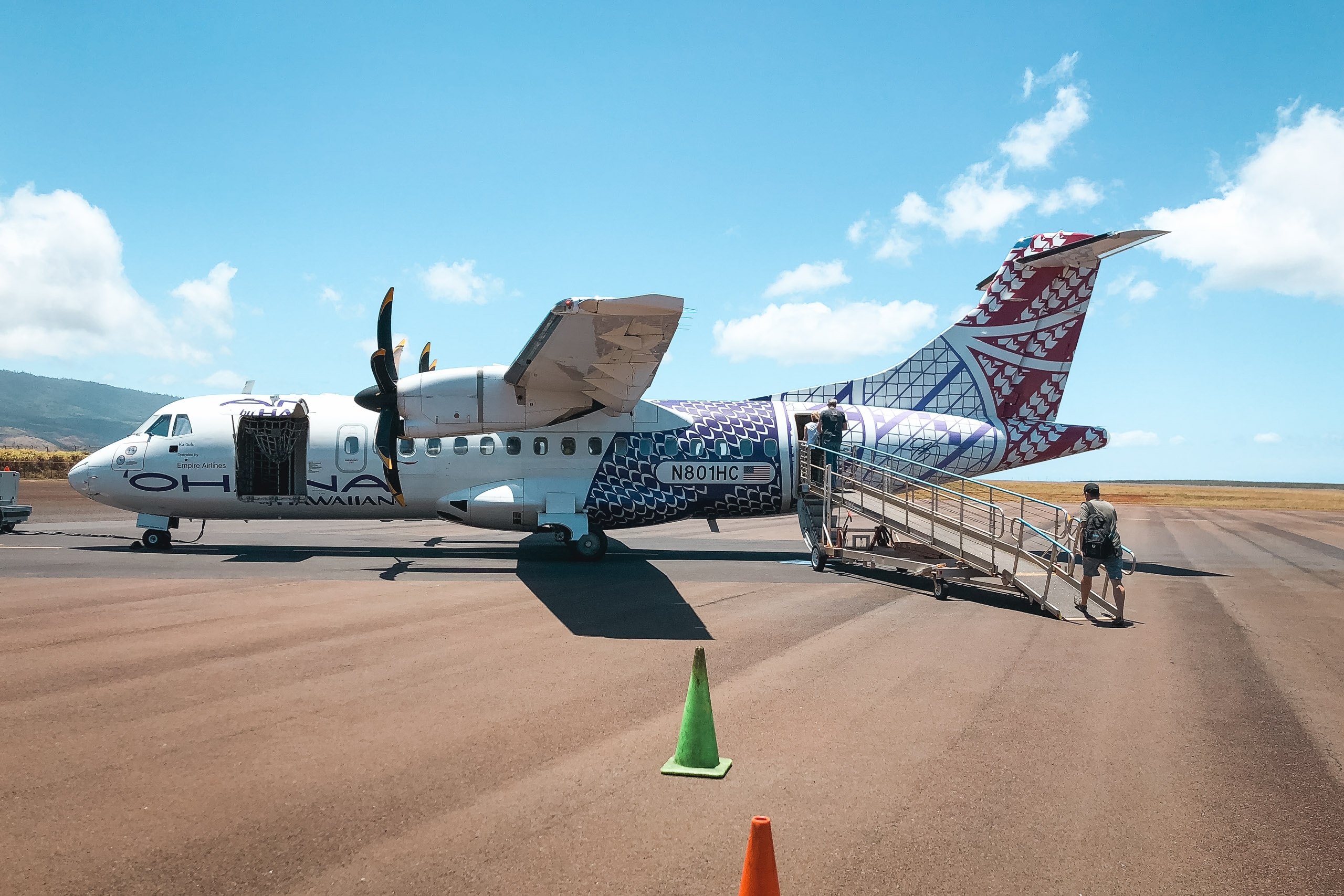 A twin-engine turboprop aircraft with decorative tail livery sits on a remote runway under clear blue sky, with boarding stairs and ground personnel visible beside it