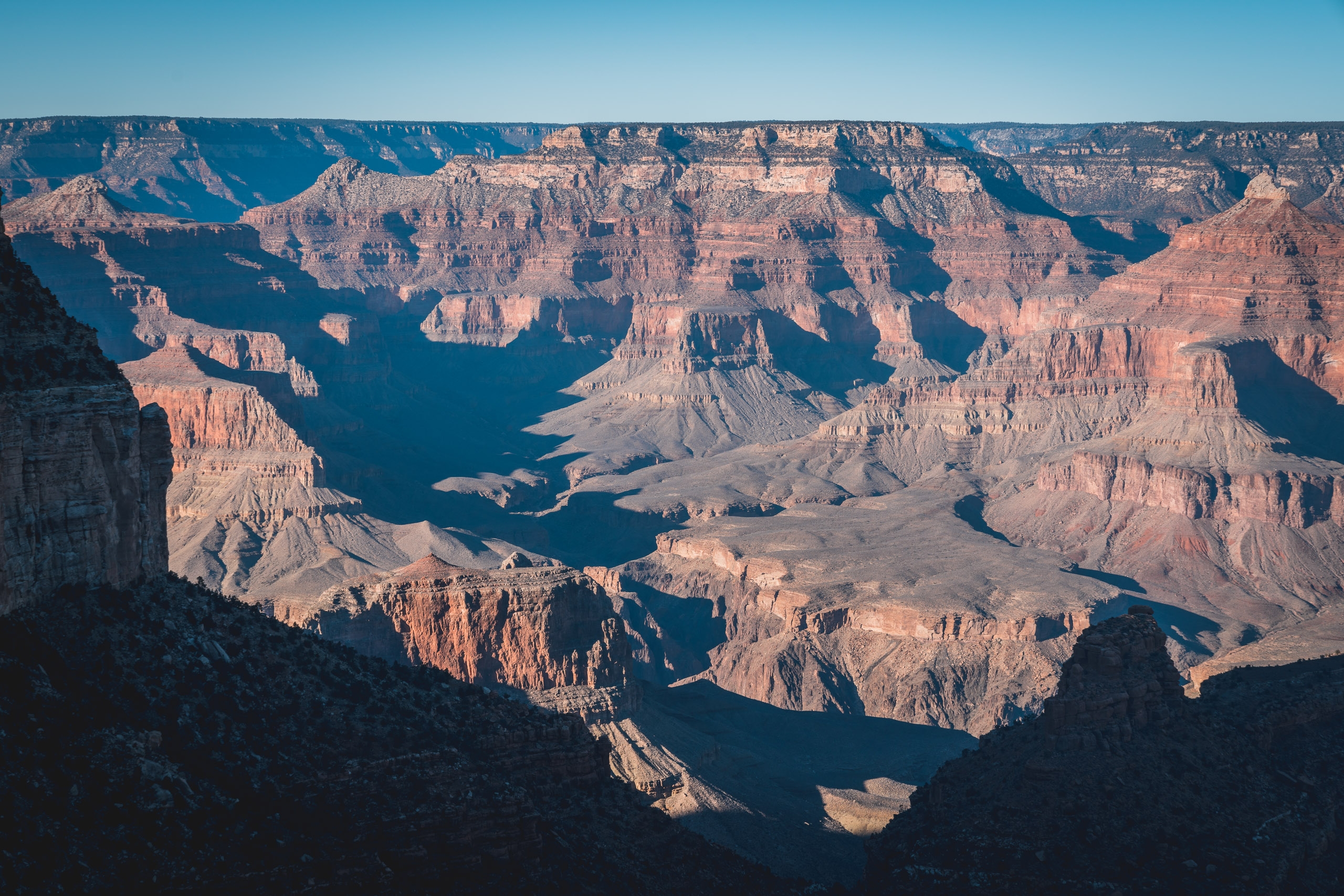 The Colorado River winds through layered red rock formations and deep shadows within the vast Grand Canyon under clear blue sky