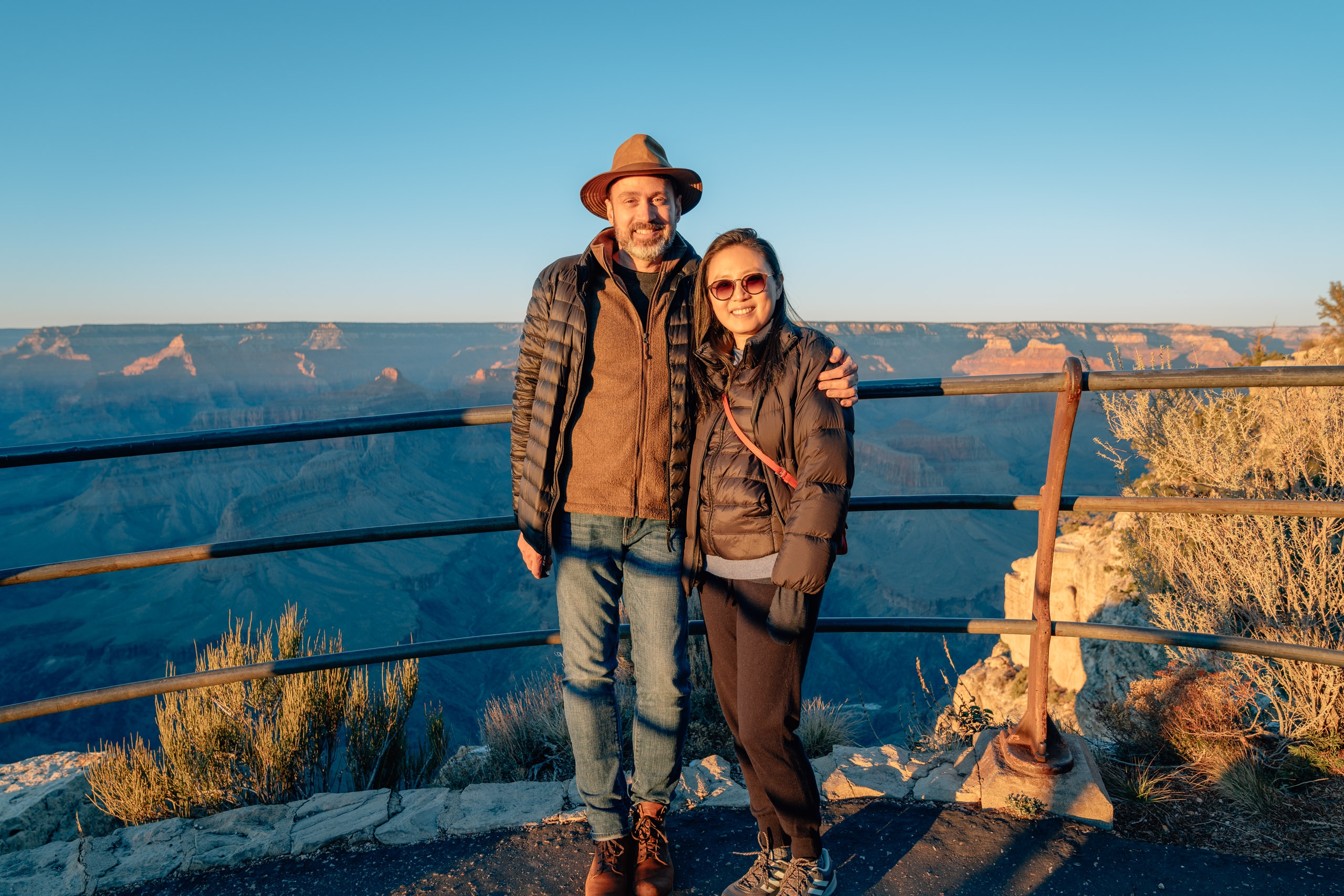 A man and woman stand arm-in-arm at a wooden railing overlooking the Grand Canyon, bathed in golden afternoon sunlight with layered canyon walls stretching across the background