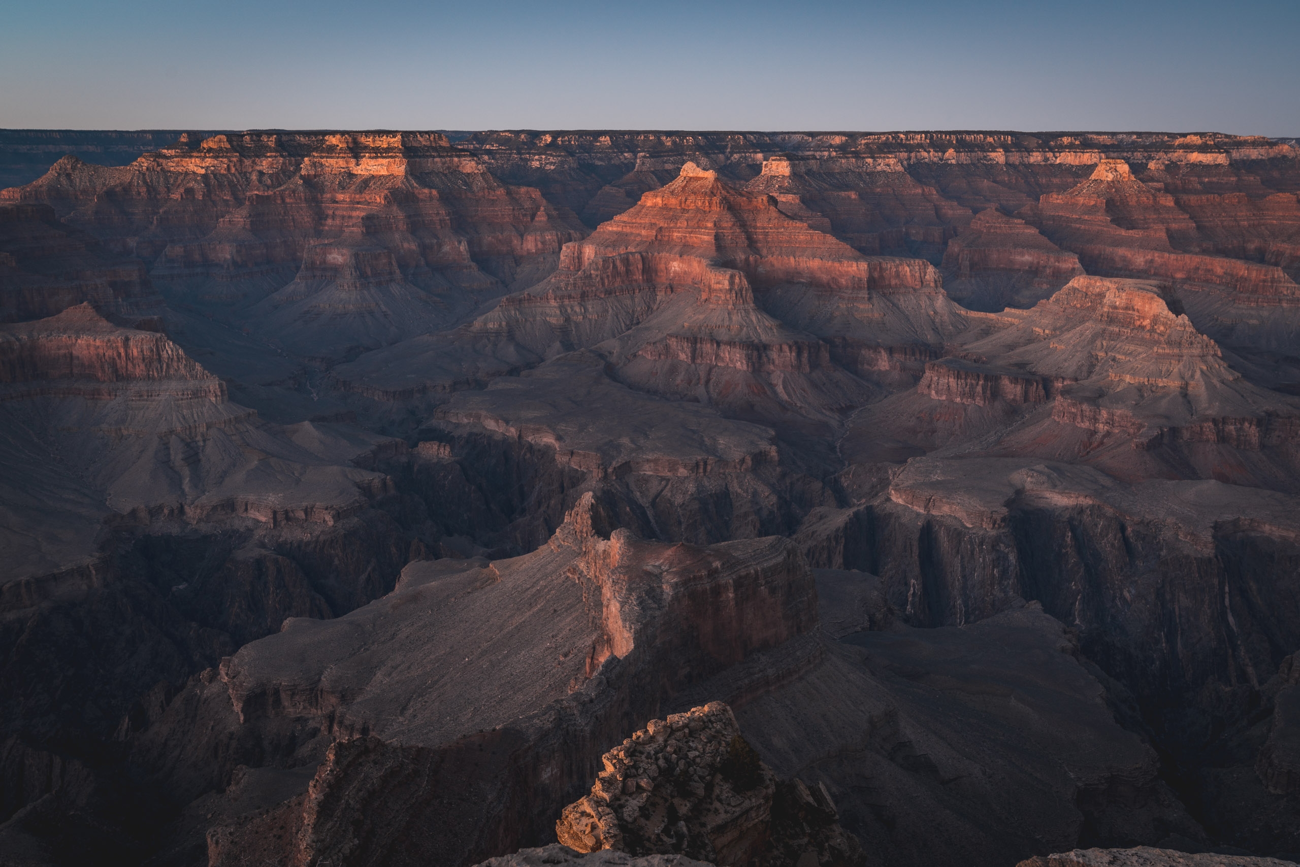 Golden sunlight illuminates the layered rock formations and deep shadows of the Grand Canyon, revealing its vast expanse of stratified cliffs and plateaus under clear sky