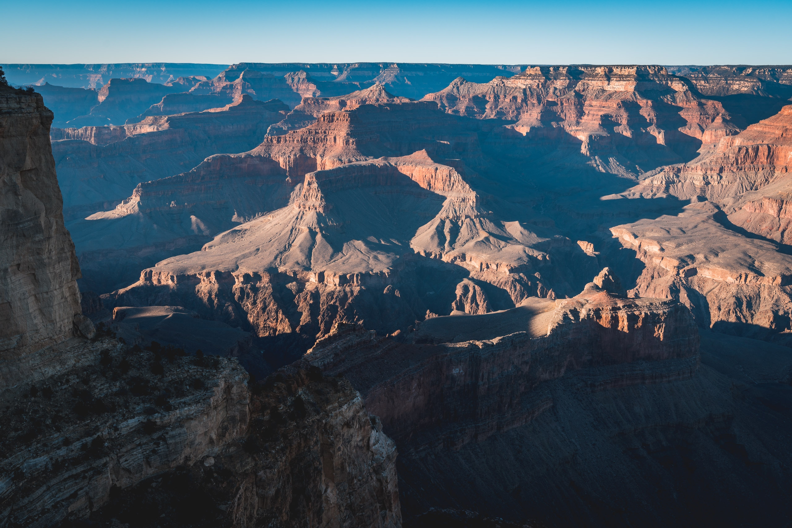 Layered red rock formations and deep shadows define the Grand Canyon's vast expanse under clear blue sky, with warm sunlight illuminating distant buttes and canyon walls