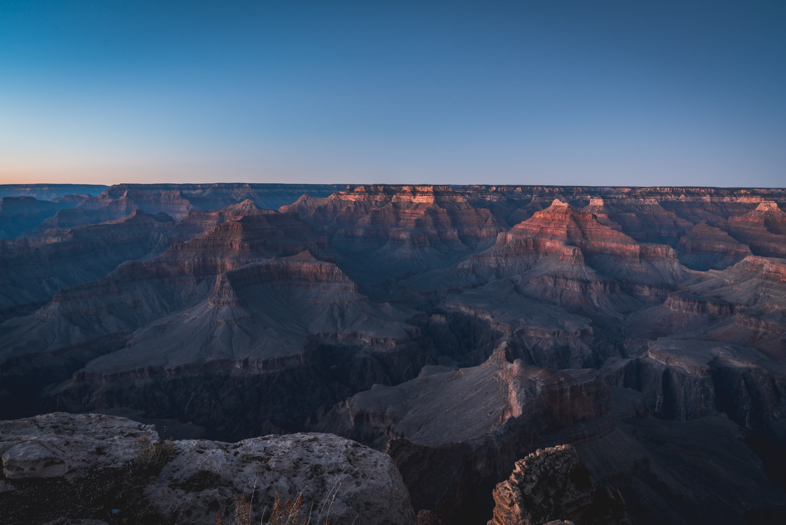 Layered rock formations of the Grand Canyon glow with warm orange light during sunset, their shadows deepening across the vast canyon under a clear blue sky