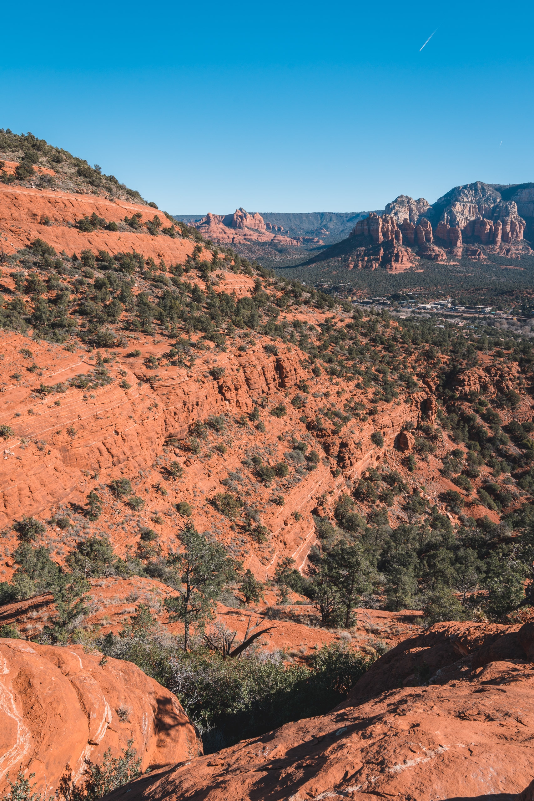 Red rock formations and scattered desert vegetation create a layered landscape in Sedona, Arizona, beneath a clear blue sky with a distant contrail