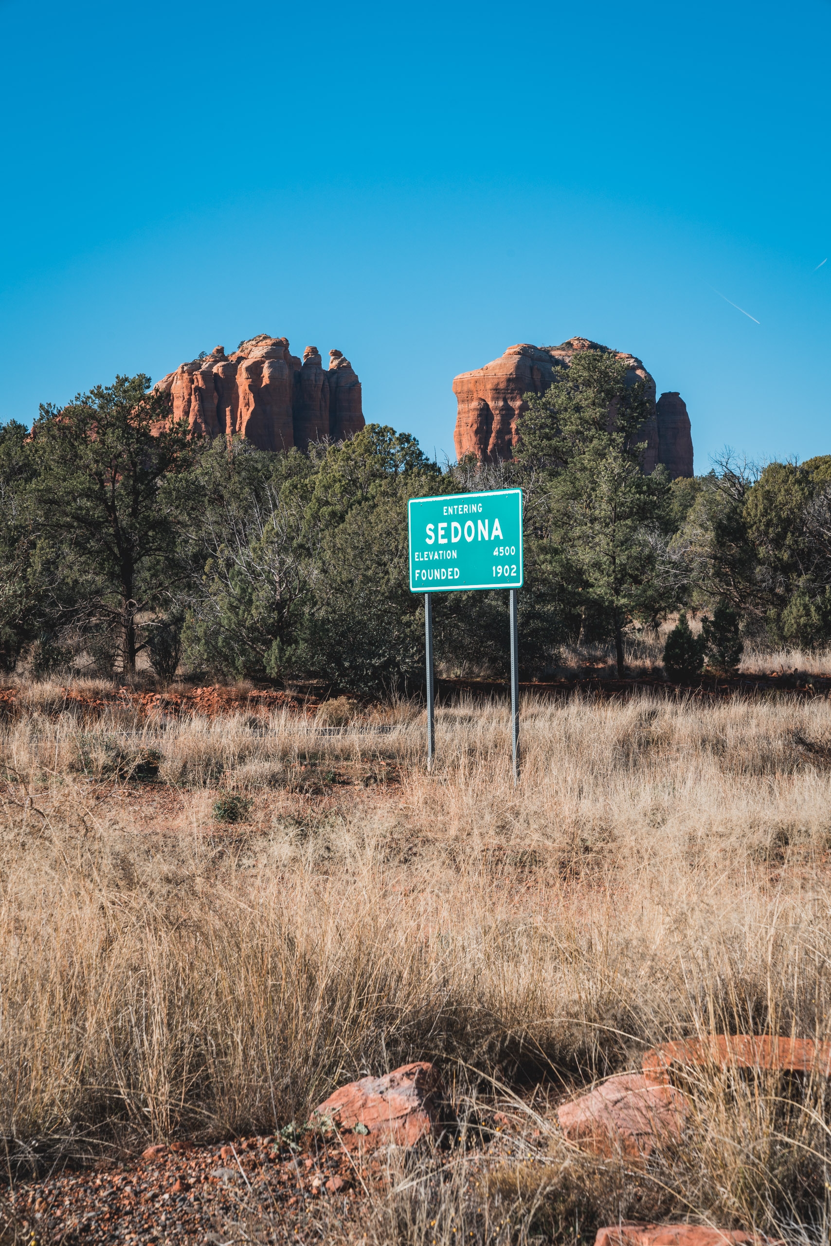 A green Sedona sign stands in dry desert grassland with red rock formations rising prominently in the clear blue sky behind it