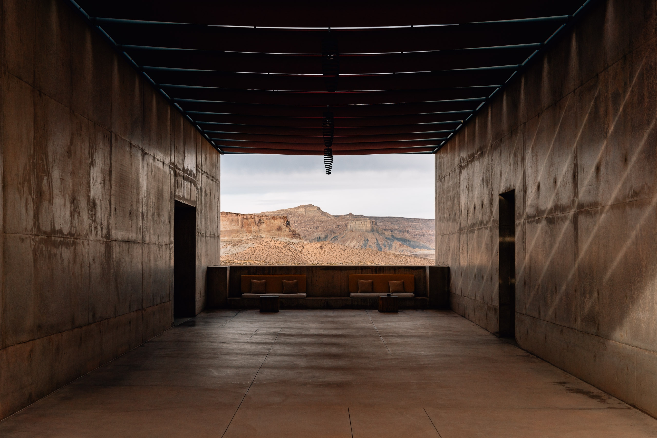 A concrete viewing pavilion frames a distant desert mountain through its open aperture, with wooden benches centered on the polished floor beneath linear ceiling beams