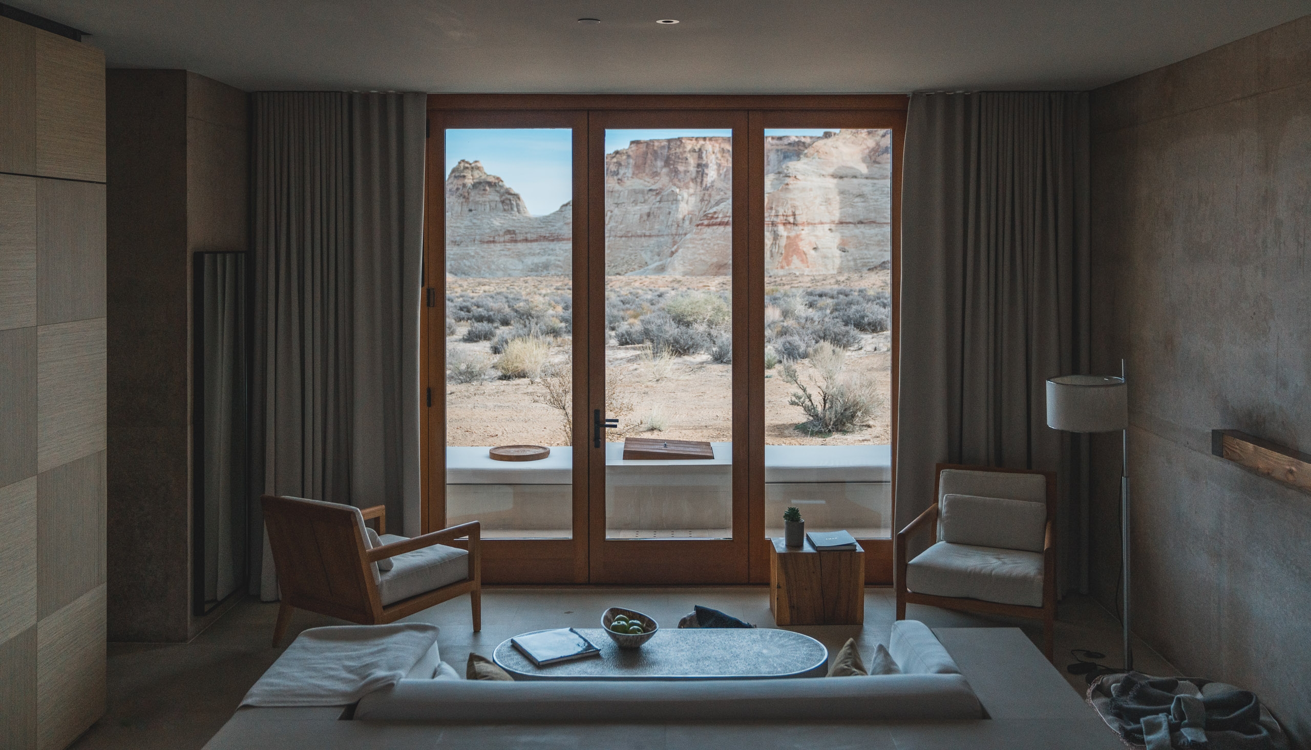 A minimalist desert resort room features two mid-century chairs facing expansive glass doors that frame eroded canyon walls and sparse vegetation under bright southwestern sunlight