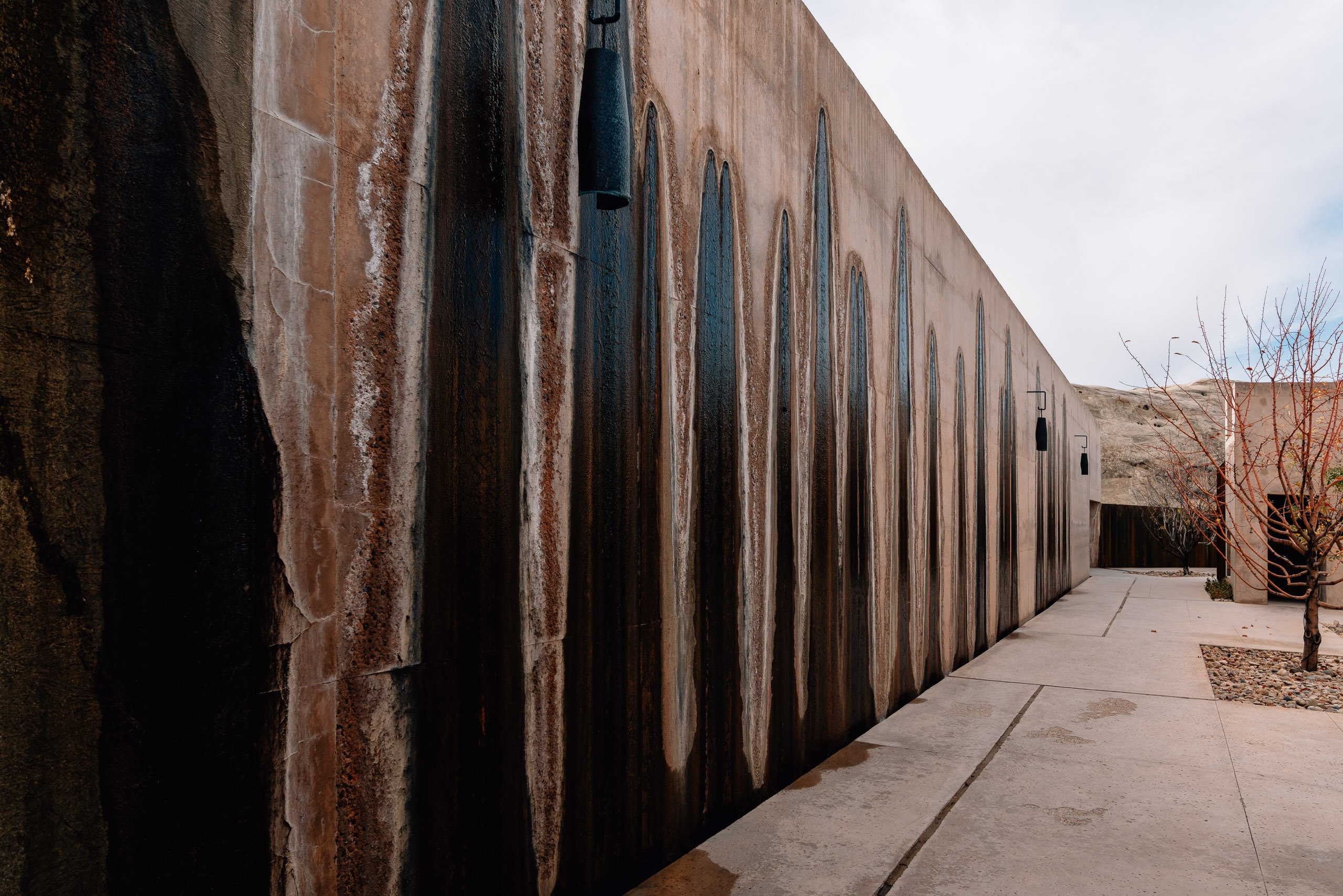 A weathered corrugated metal wall with deep vertical ridges lines a concrete pathway beneath overcast skies, creating strong geometric shadows and textural contrast in this desert resort courtyard