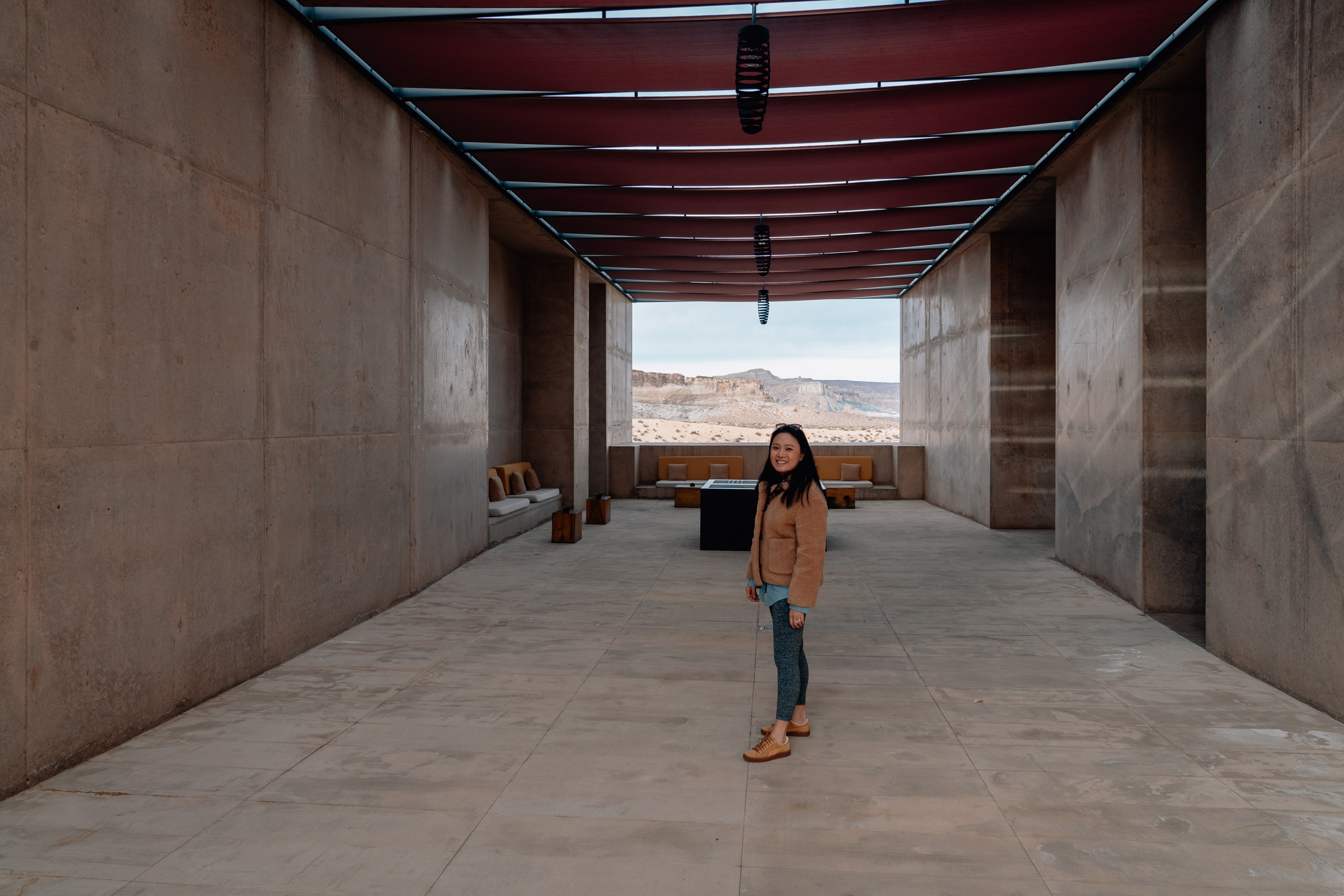 Woman in caramel jacket stands centered in a minimalist concrete corridor with burgundy ceiling beams, framing distant desert mountains through the open end