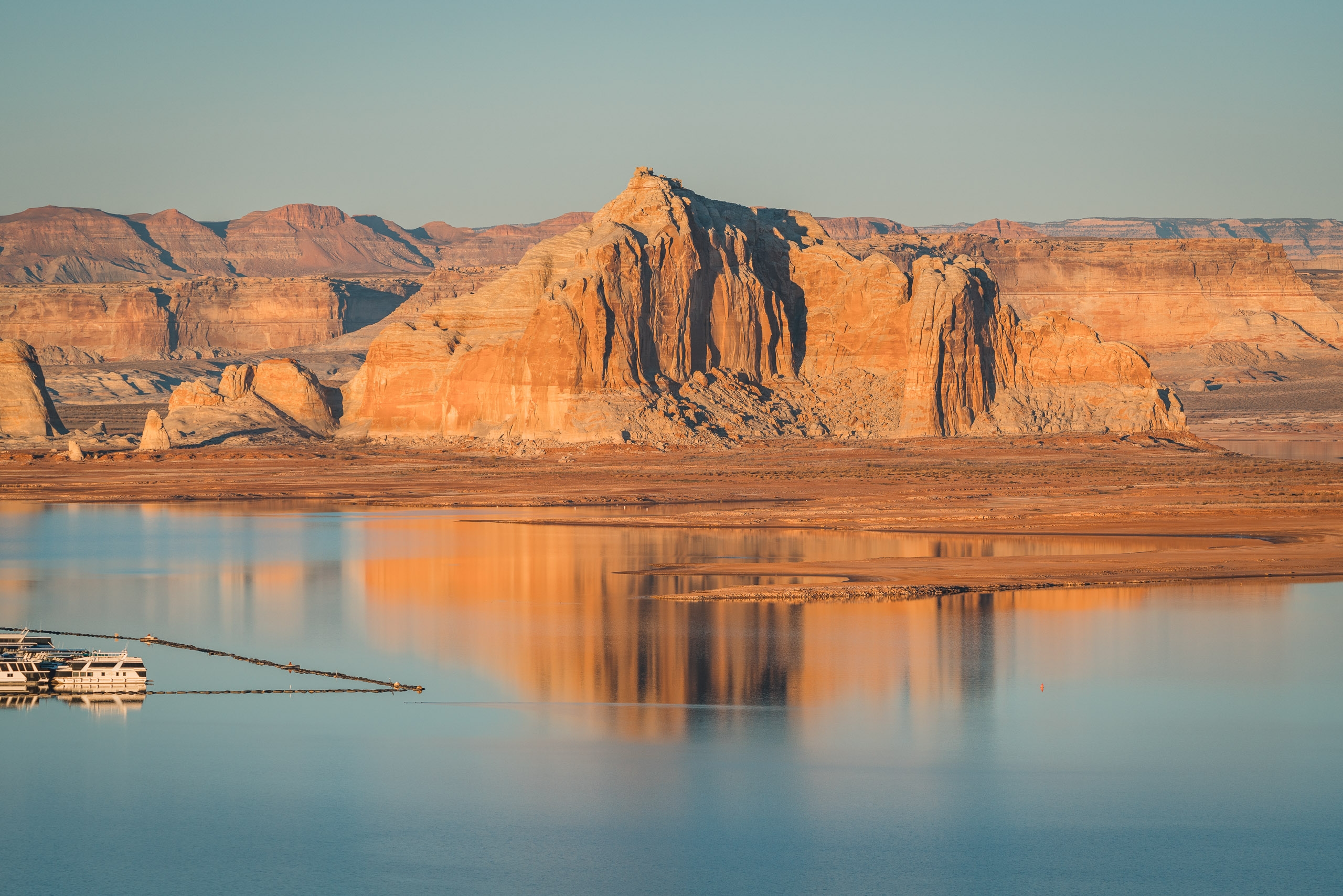 Golden-hued rock formations rise majestically above the still, mirror-like waters of Lake Powell under clear desert light at dawn or dusk