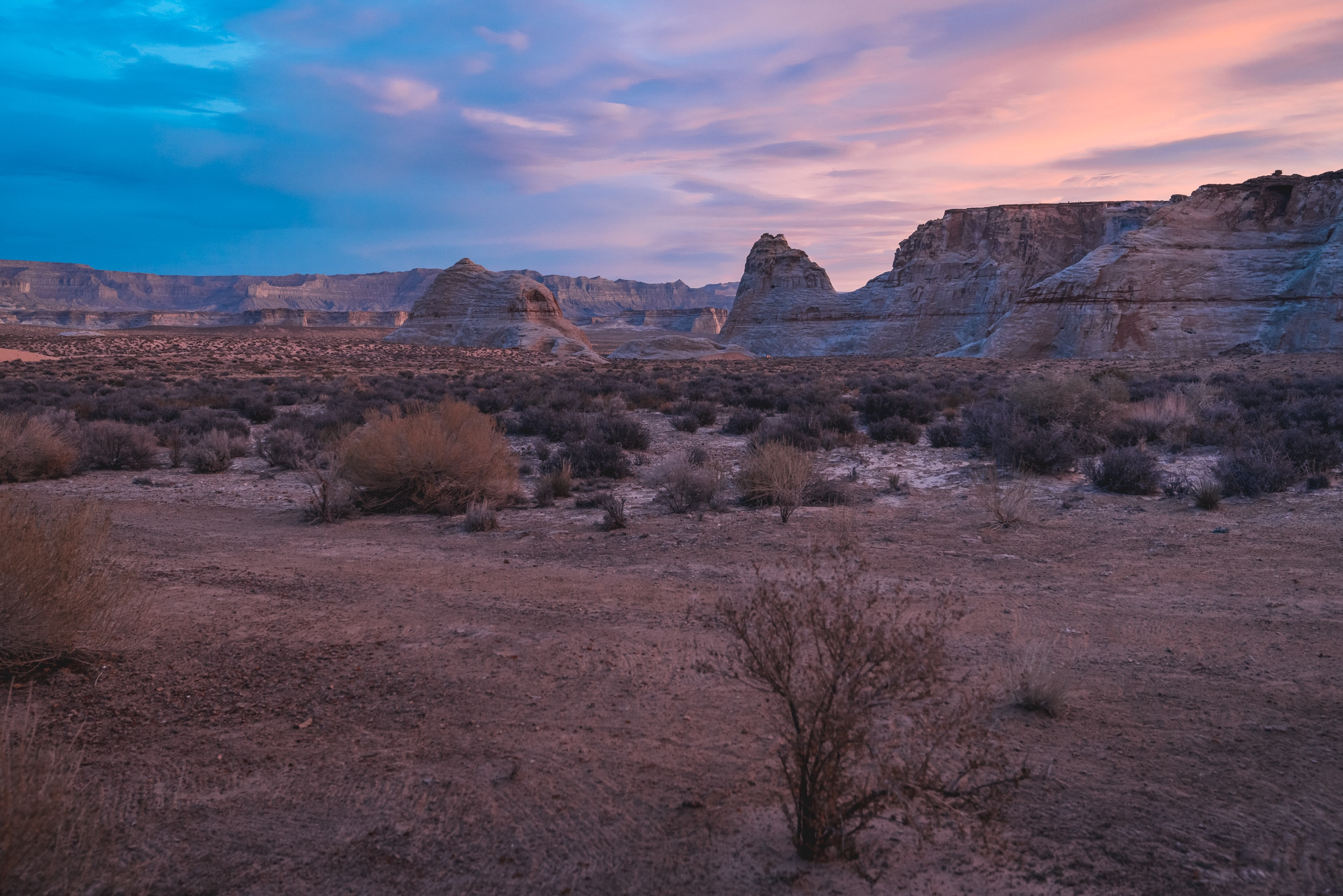 Desert landscape at dusk with layered rock formations and sparse vegetation under a pink and blue twilight sky