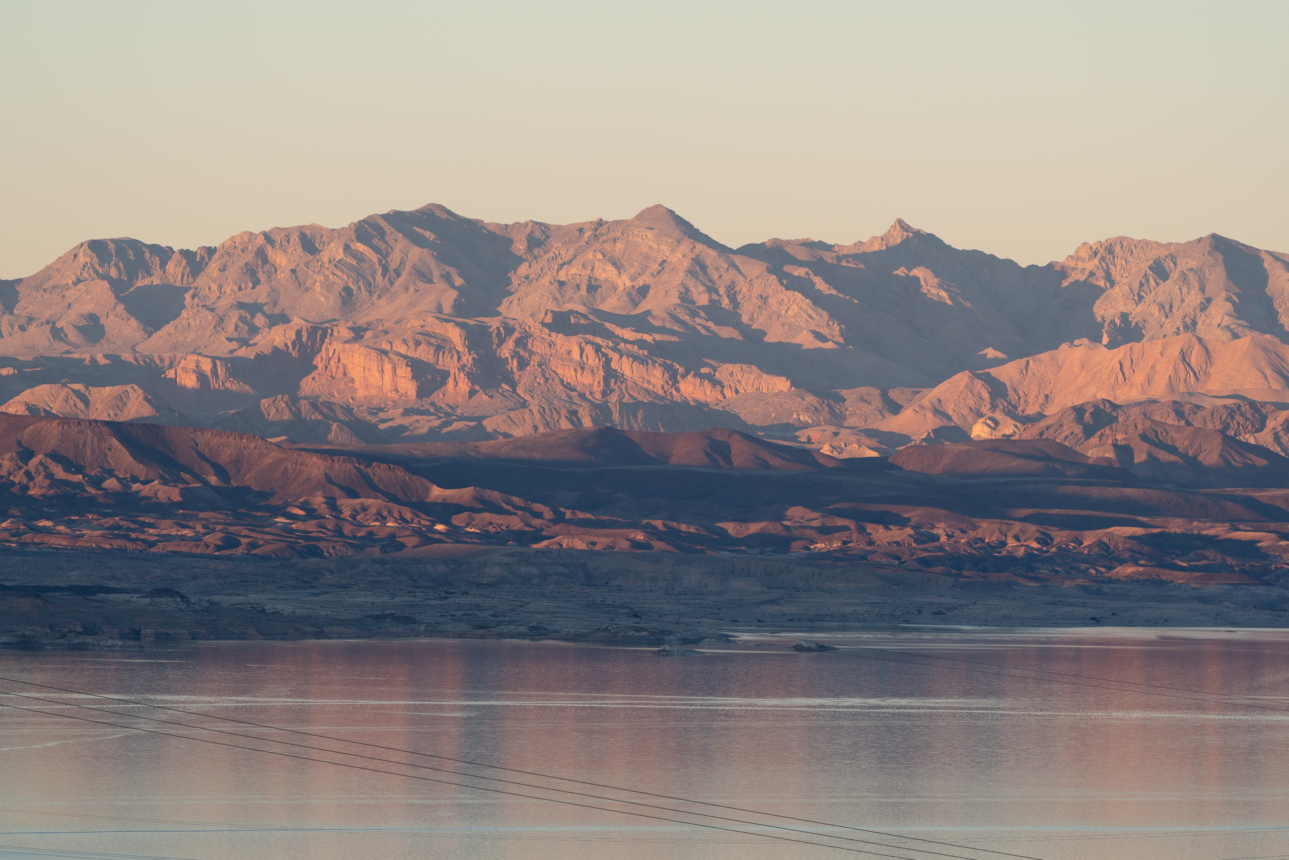 Rugged desert mountains with warm golden light reflect across still water in the foreground, with power lines stretching across the calm lake surface at dusk
