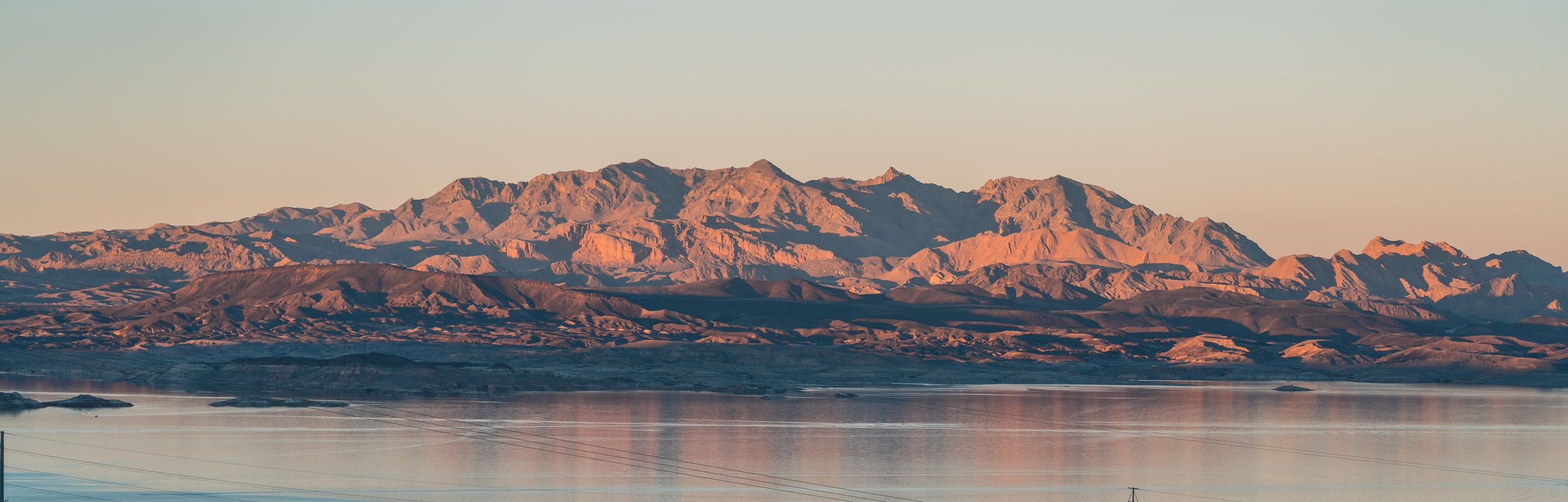 Arid mountains with rust-colored rock formations rise behind a calm lake reflecting warm sunset light, creating a serene desert landscape